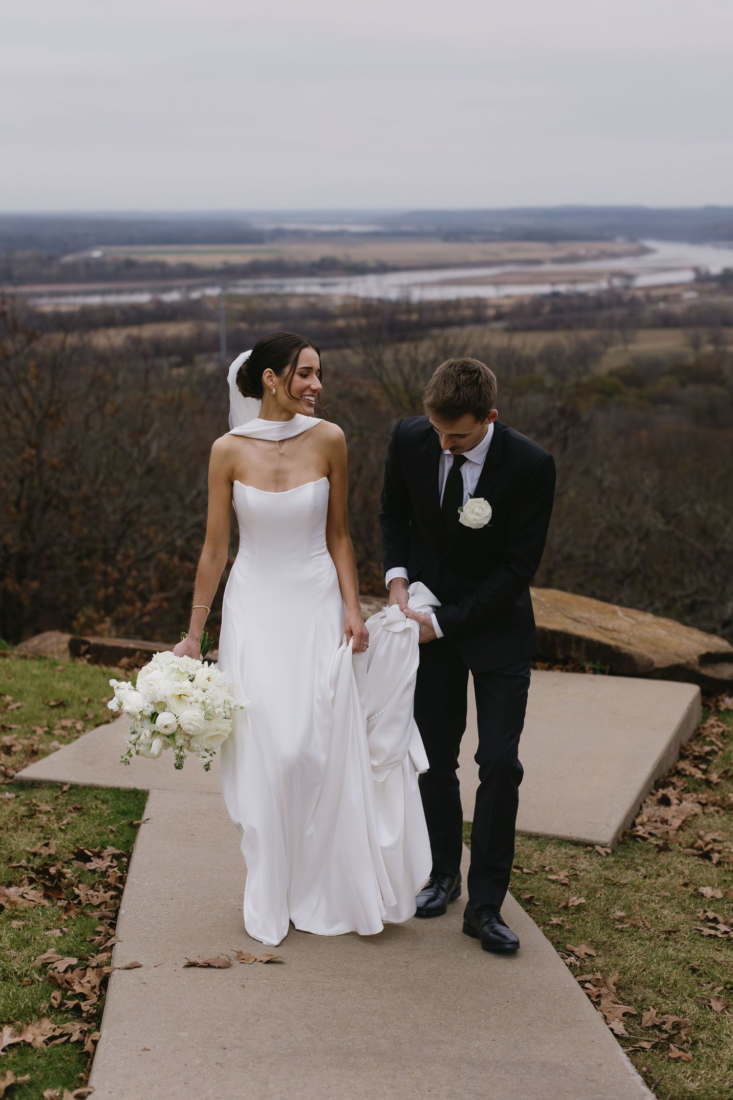 Wedding couple walking down concrete path with scenic overlook in background.