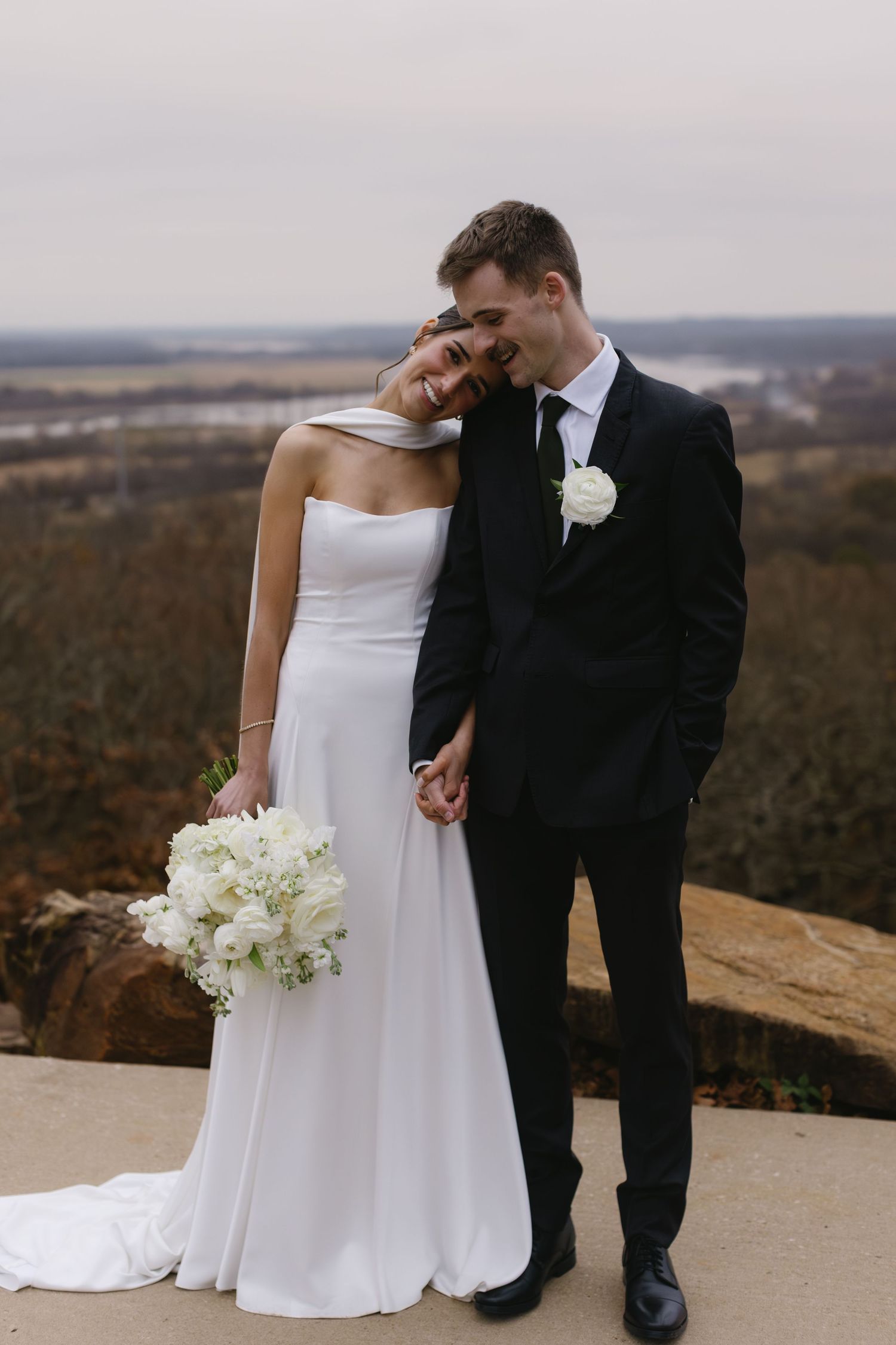 Wedding couple shares intimate moment on scenic overlook with rolling hills in background.