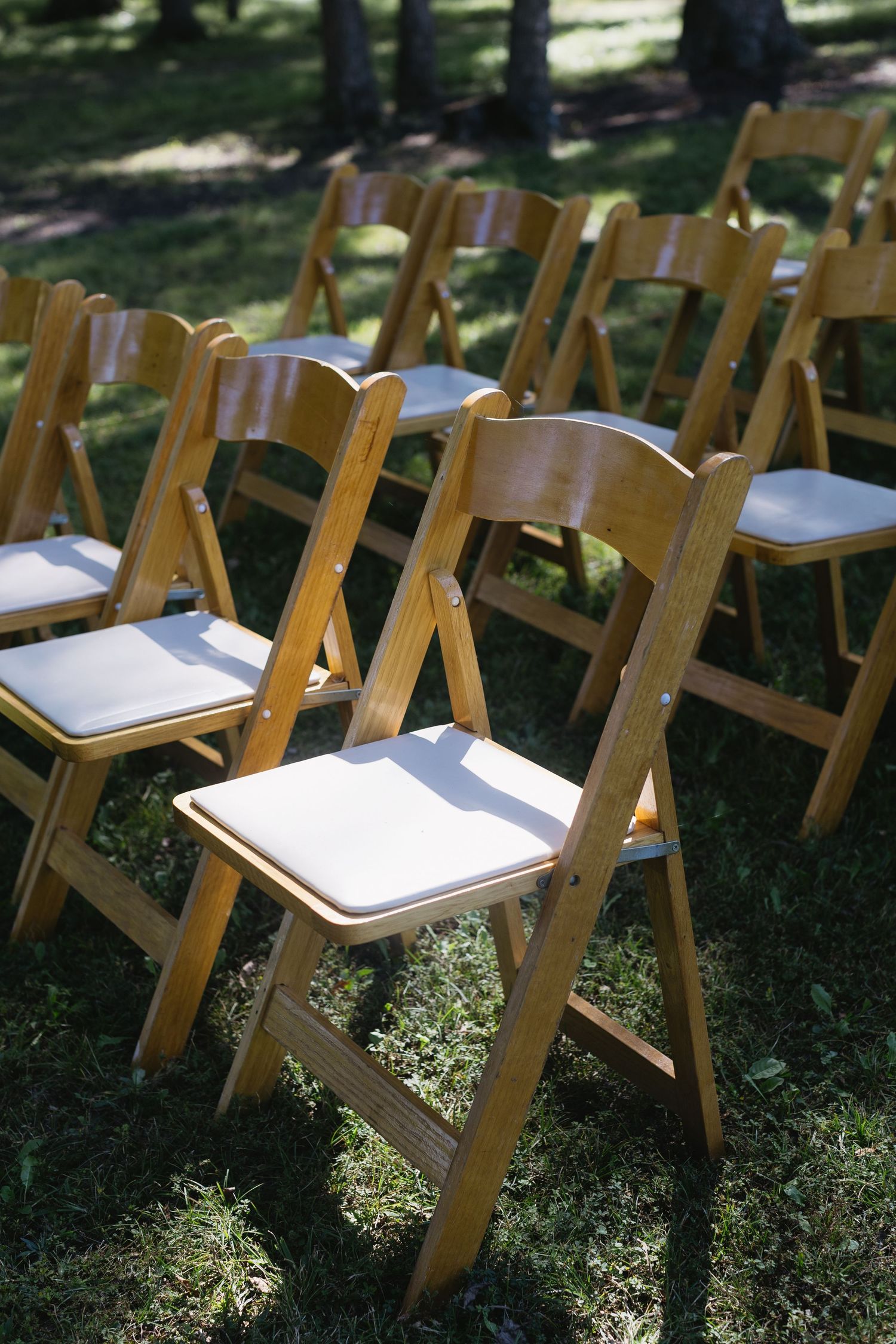 Wooden folding chairs with white cushions arranged in rows on grass for an outdoor ceremony.