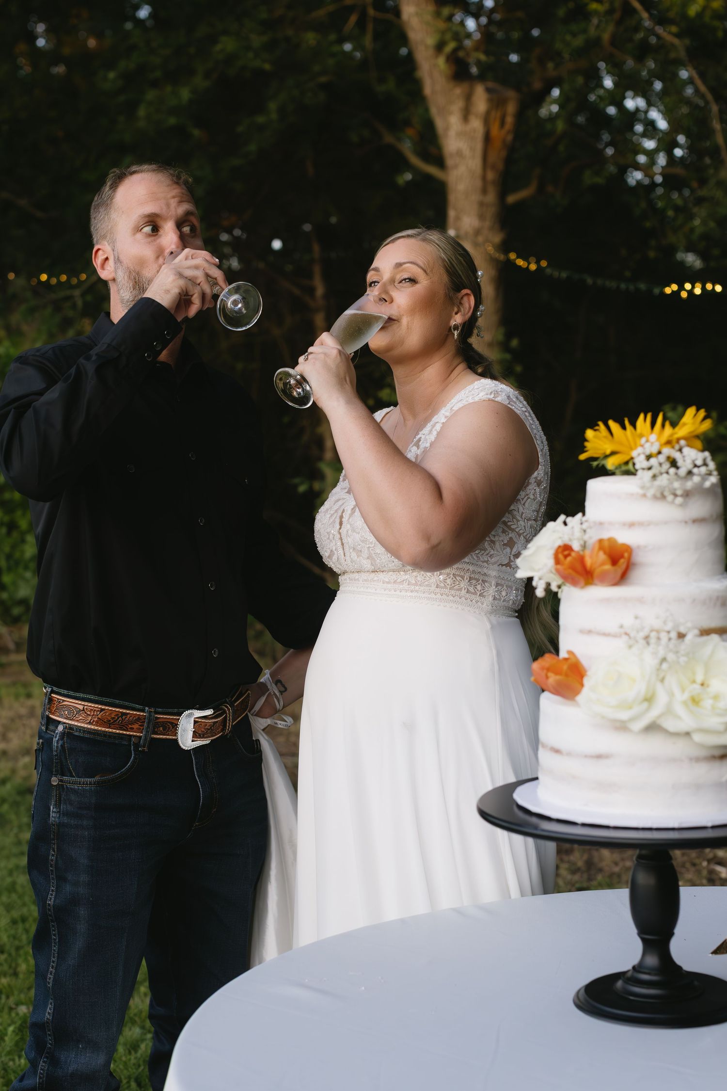 A couple toasts with champagne glasses next to their white wedding cake decorated with sunflowers.
