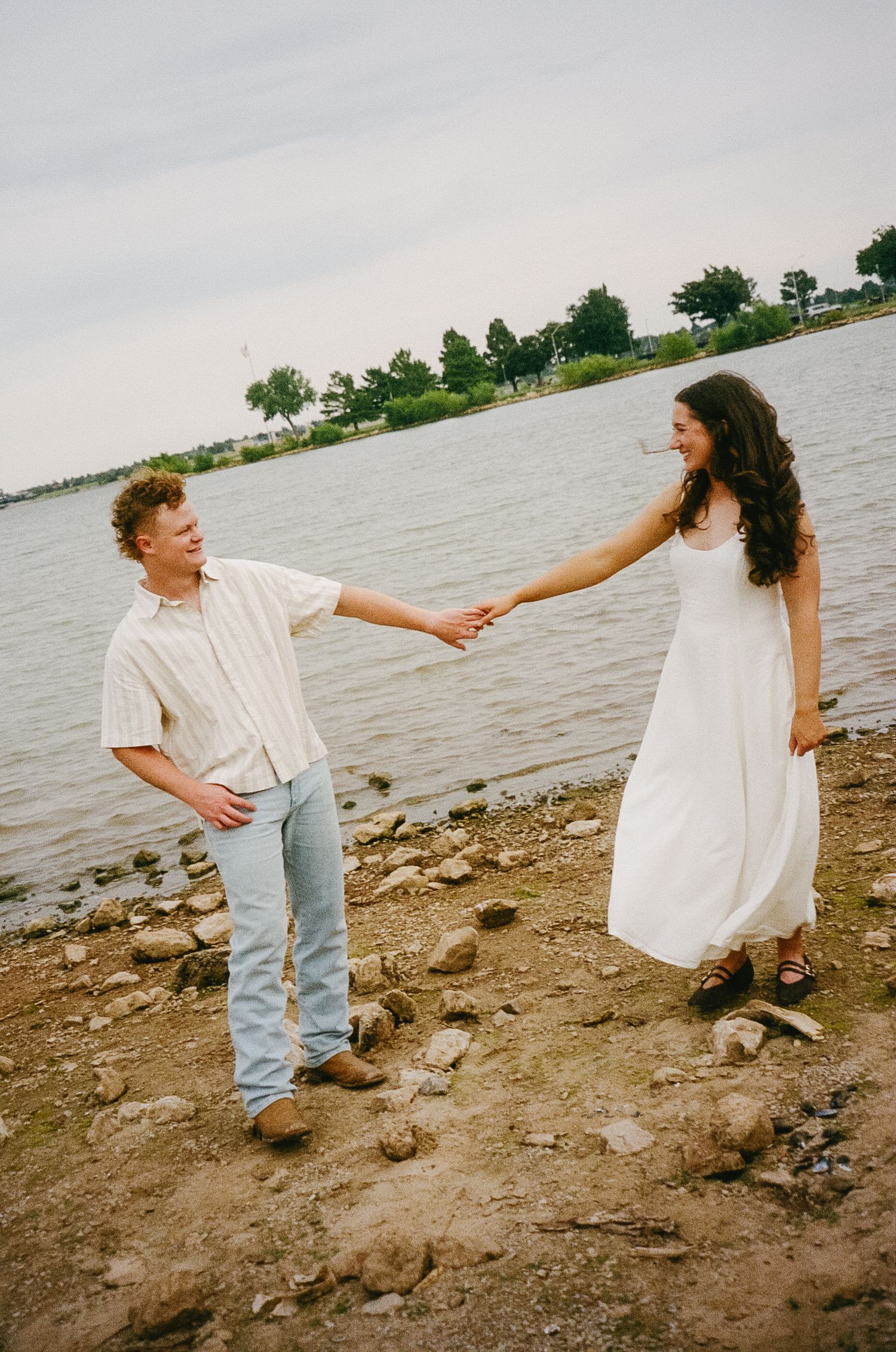 Couple in white and denim walking along a rocky shoreline holding hands near the water.