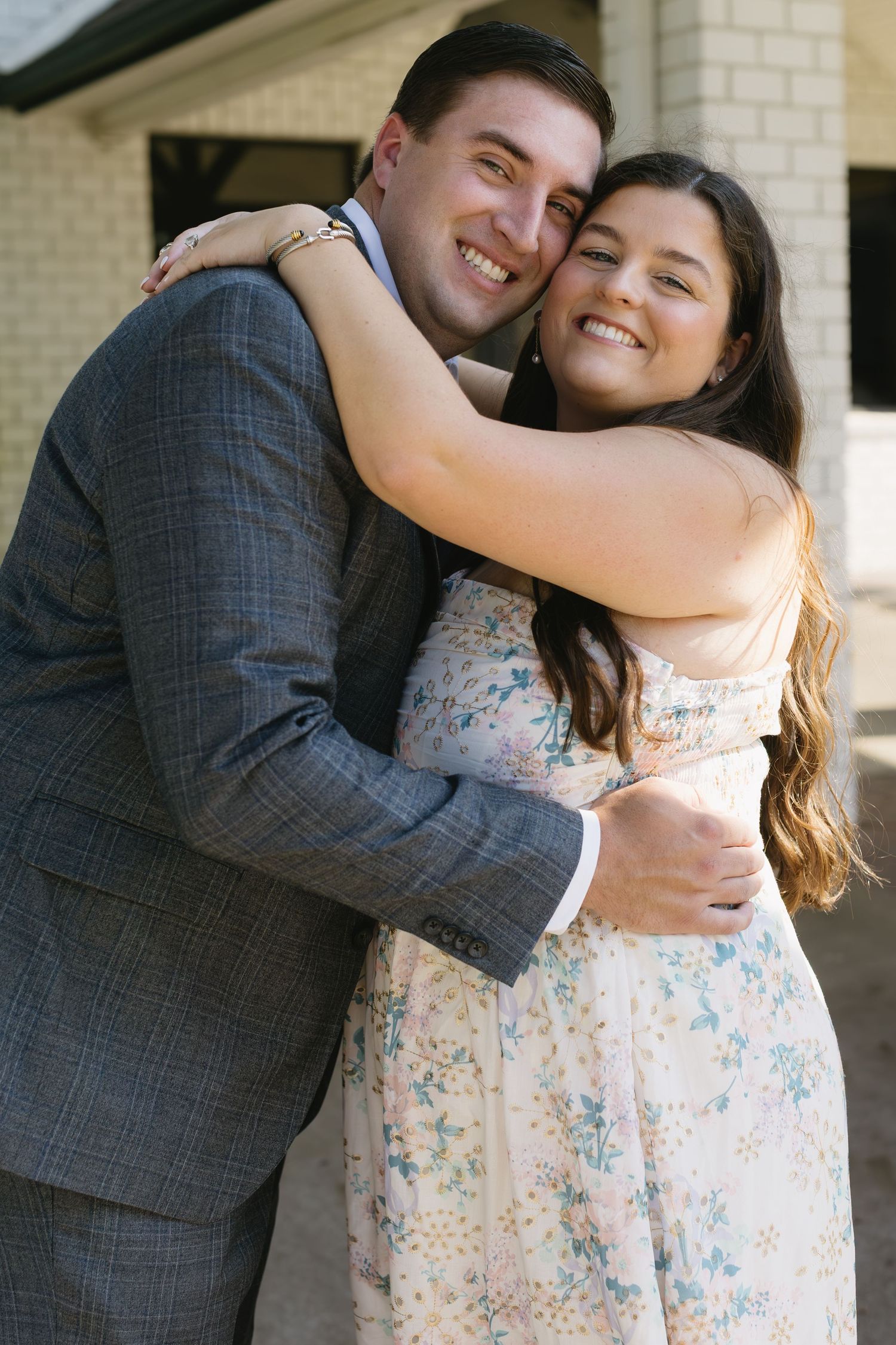 A romantic couple embraces outdoors in front of a light colored brick wall, with a floral dress and grey suit.