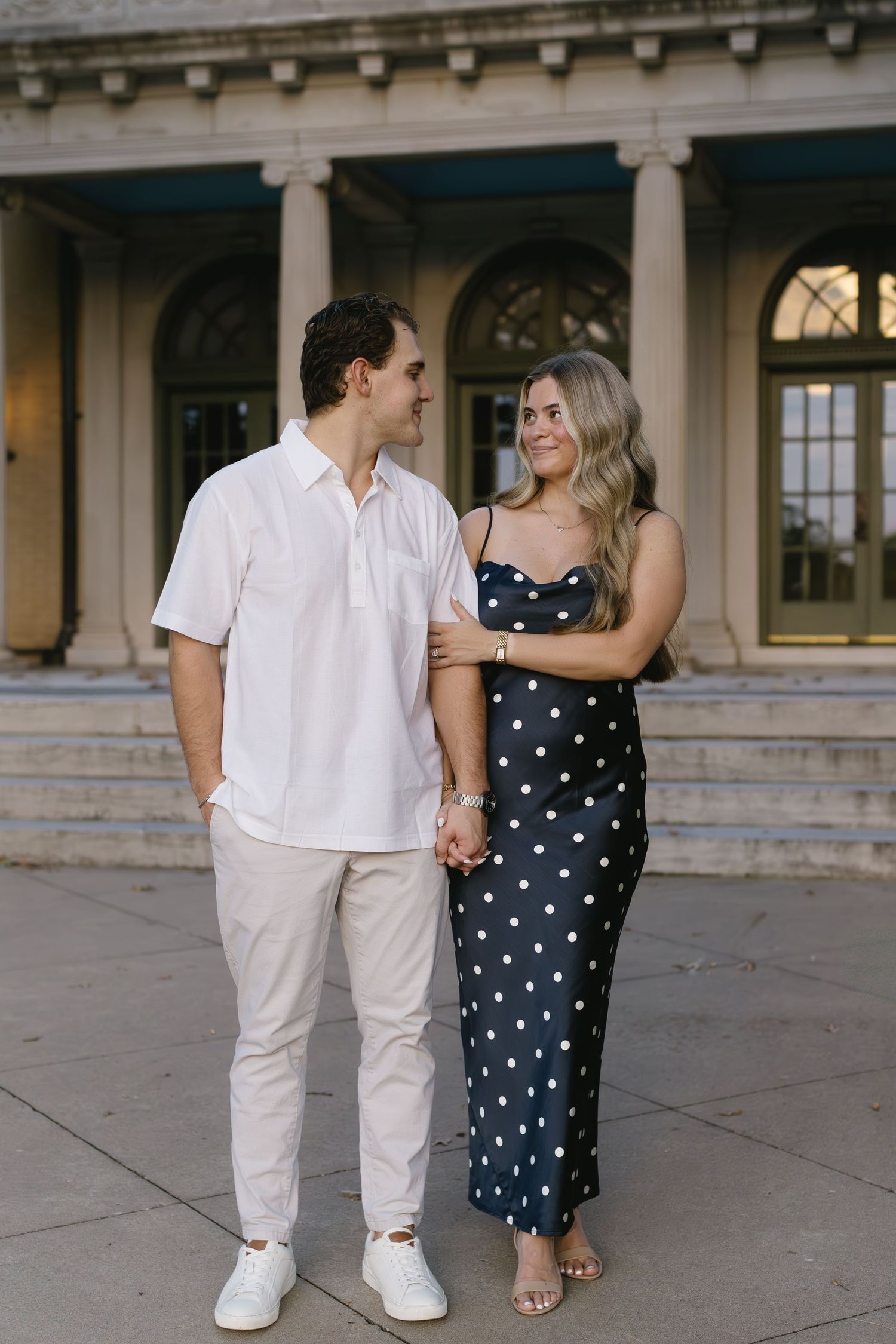 Couple strolling together in coordinated white and polka dot outfits near a historic building.