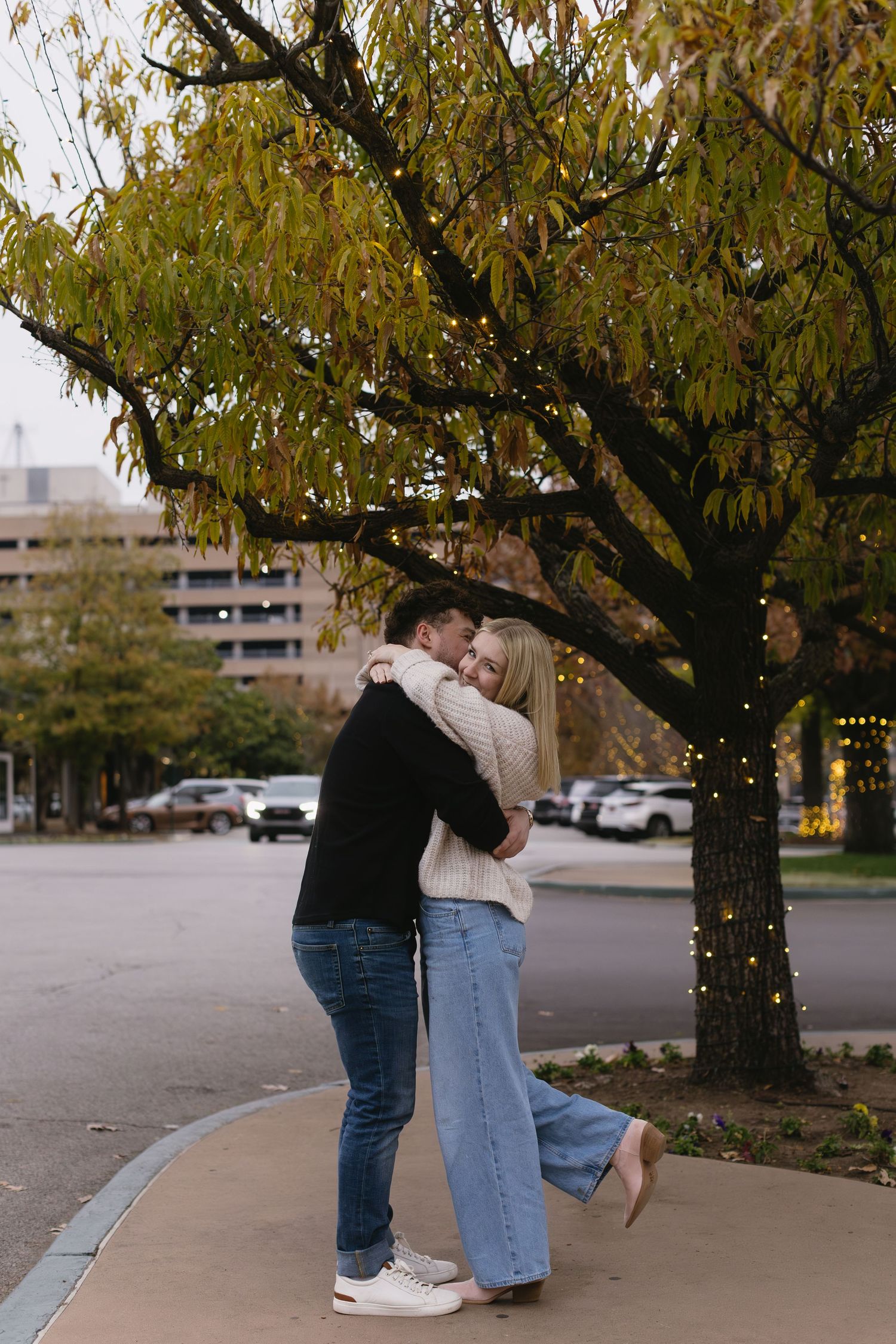 A couple shares an intimate moment under a tree with twinkling lights in an urban setting.