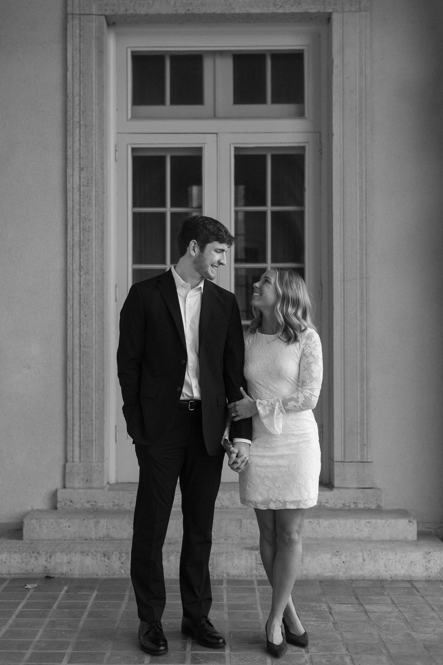 A couple in formal wear stands together in front of a large window in black and white.