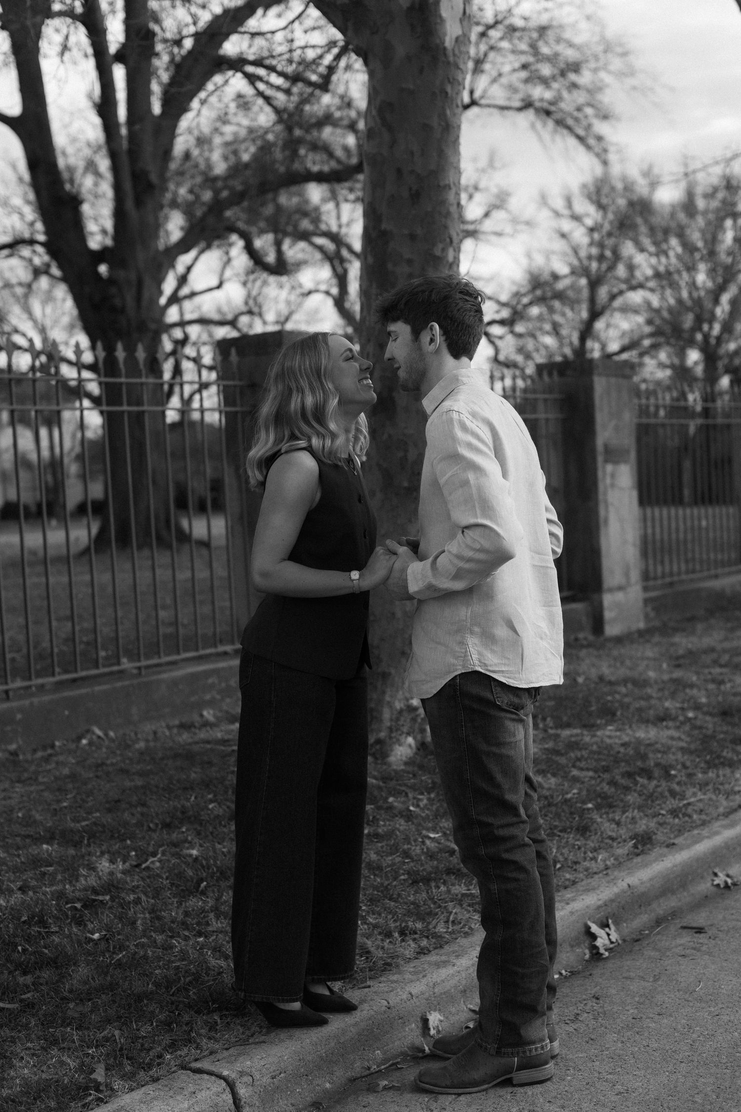A couple shares a moment together on a sidewalk near iron fencing and bare trees.