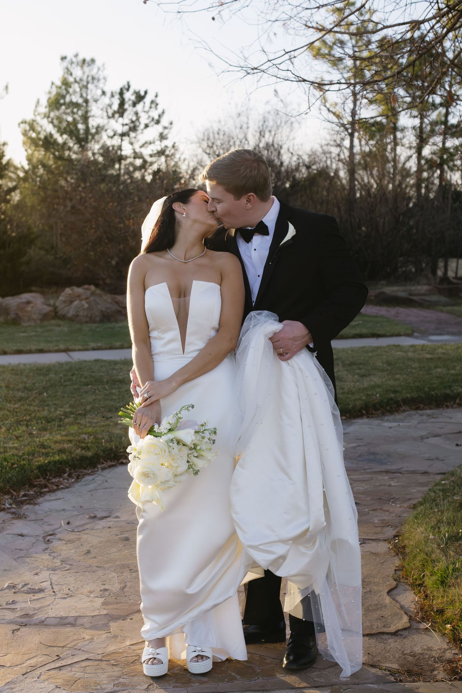 A romantic wedding couple shares a kiss on a stone pathway at sunset while the bride holds a white bouquet.