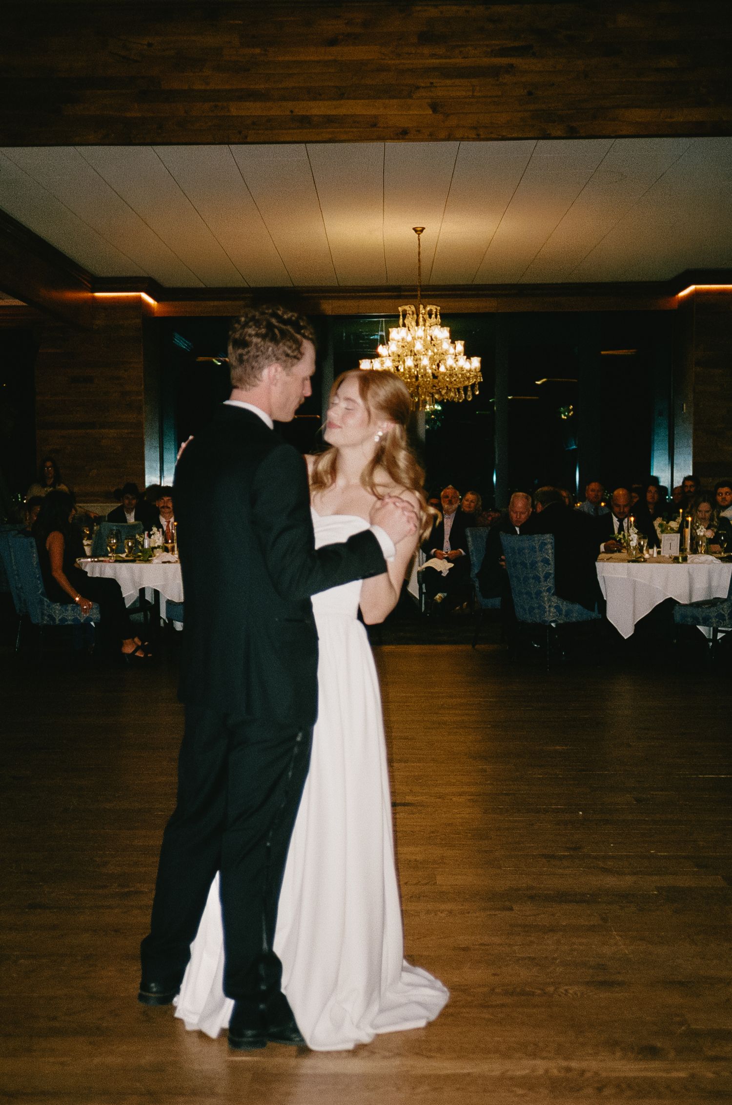 First dance in elegant reception hall with wooden floors and chandelier lighting.
