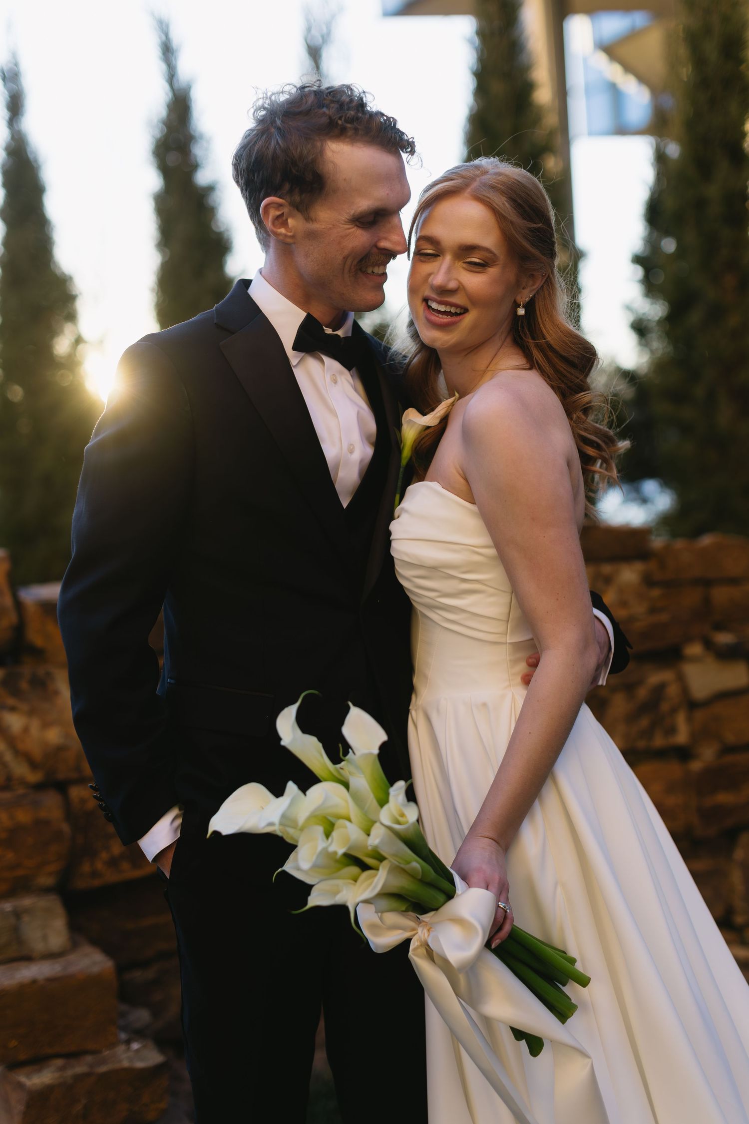 Romantic sunset wedding portrait with couple holding calla lily bouquet among cypress trees.