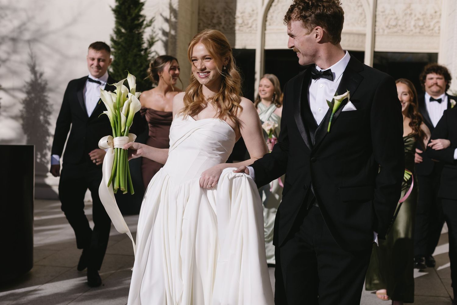Wedding party poses outdoors with bride in white dress and groomsmen in black tuxedos.