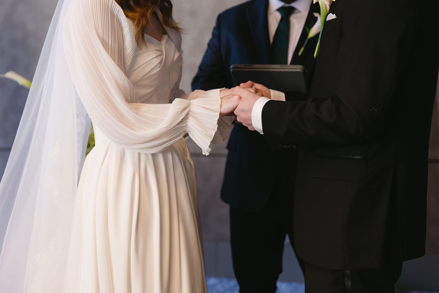 A close-up of joined hands during a wedding ceremony showing white dress and dark suit details.