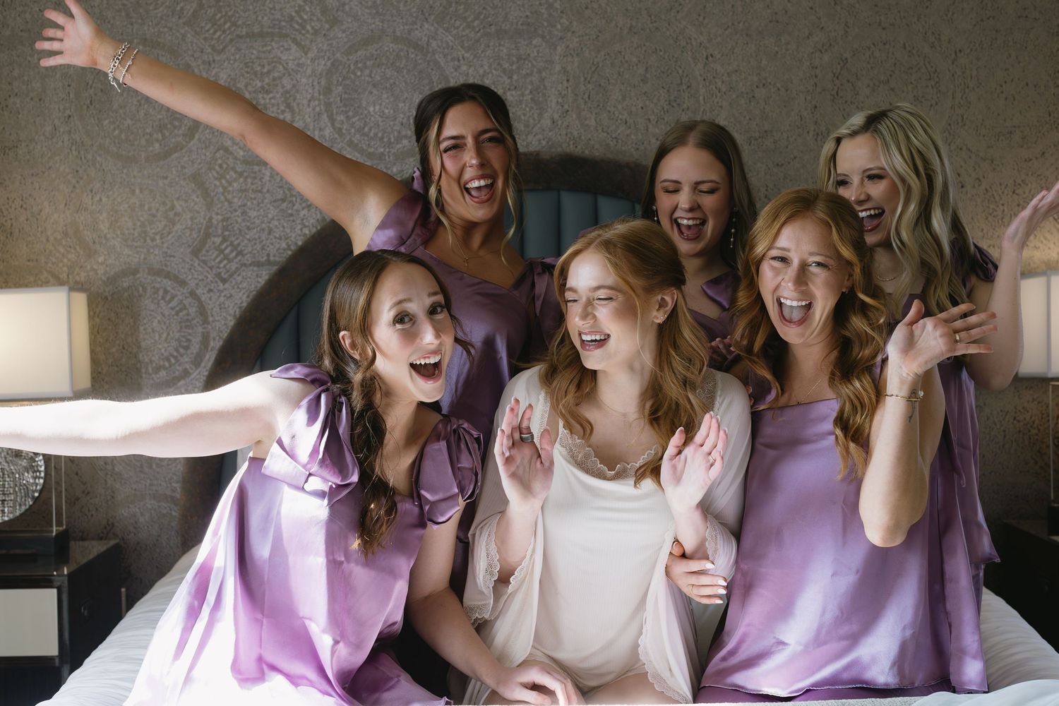 Group wearing matching purple bridesmaid dresses sitting together on a couch during wedding preparations.