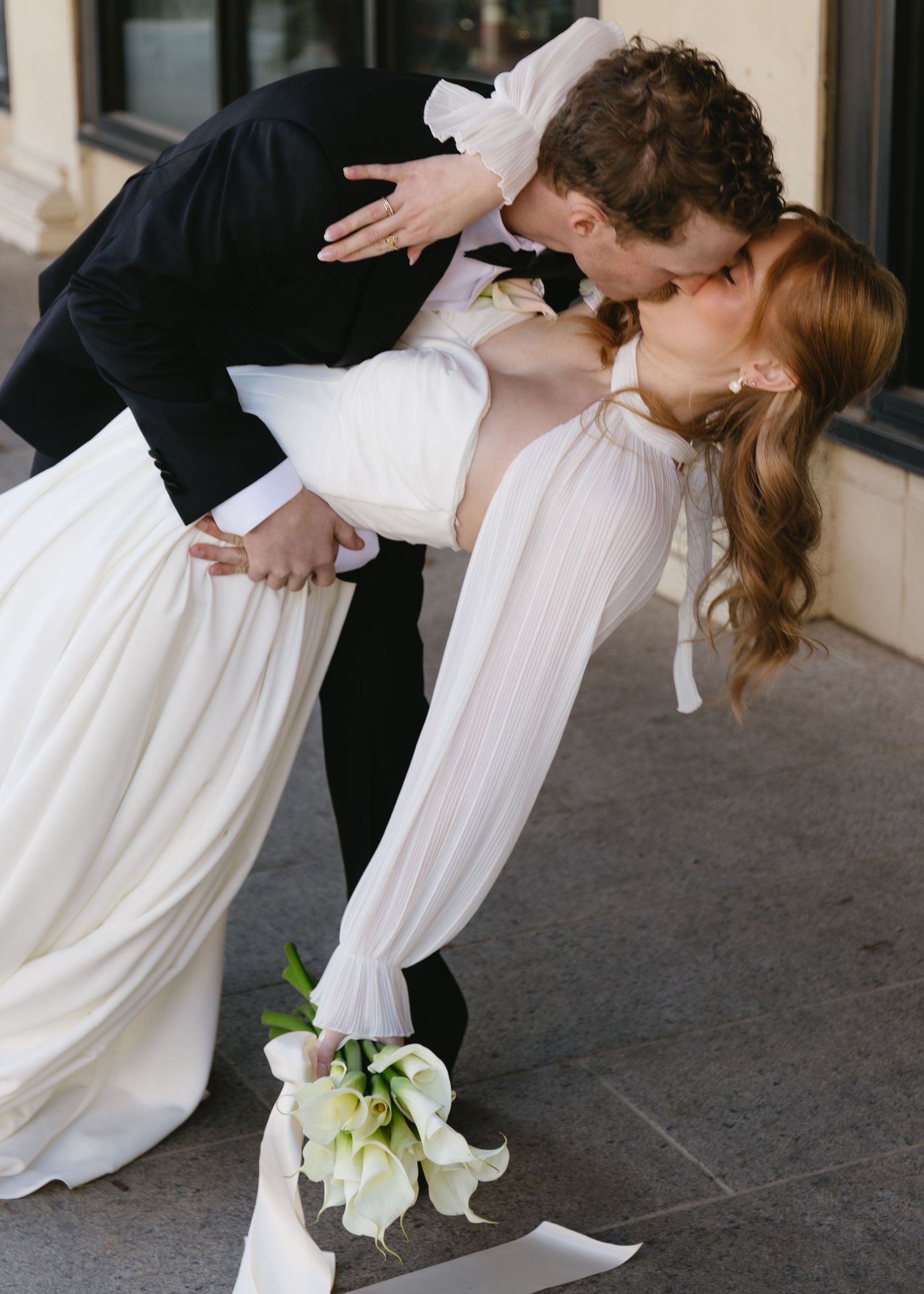 Wedding couple performs dramatic dip dance move on stone patio with calla lily bouquet.