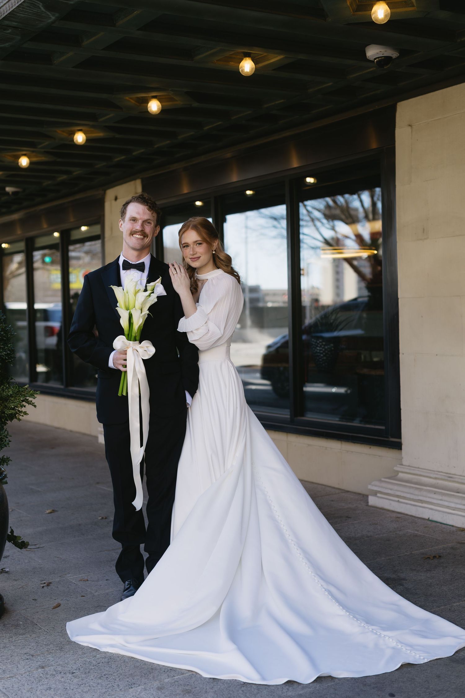 A wedding couple poses outdoors under a covered walkway with their flowing white dress.