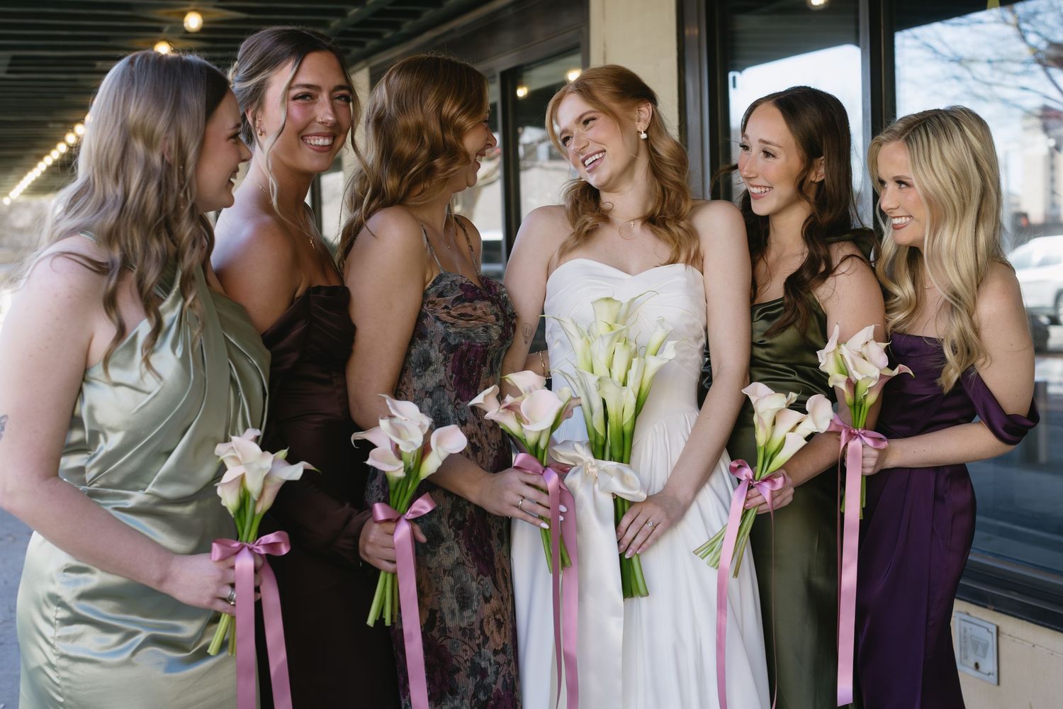 Bridesmaids in pastel dresses holding bouquets gather together at an outdoor venue.