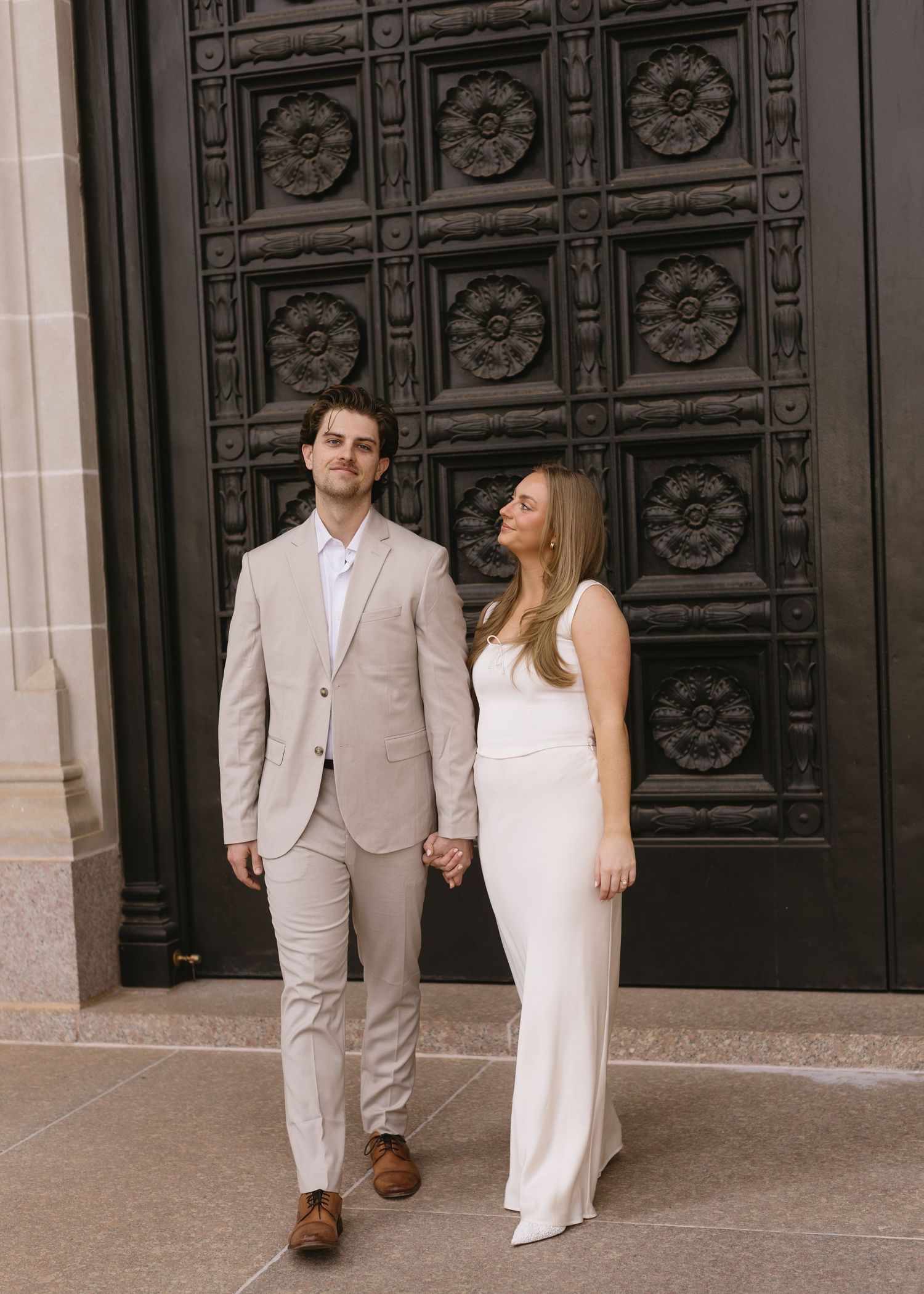 A couple in elegant wedding attire stands in front of ornate black doors at a historic building.
