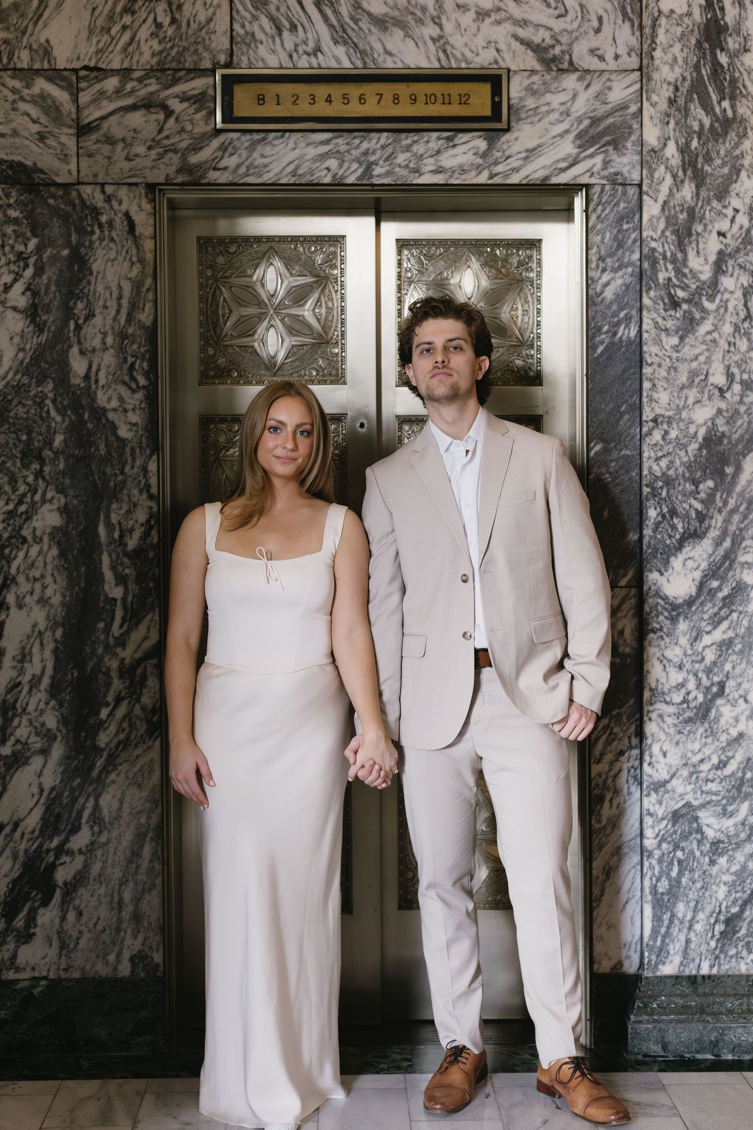 Couple in formal wear pose in front of marble walls and decorative elevator doors.