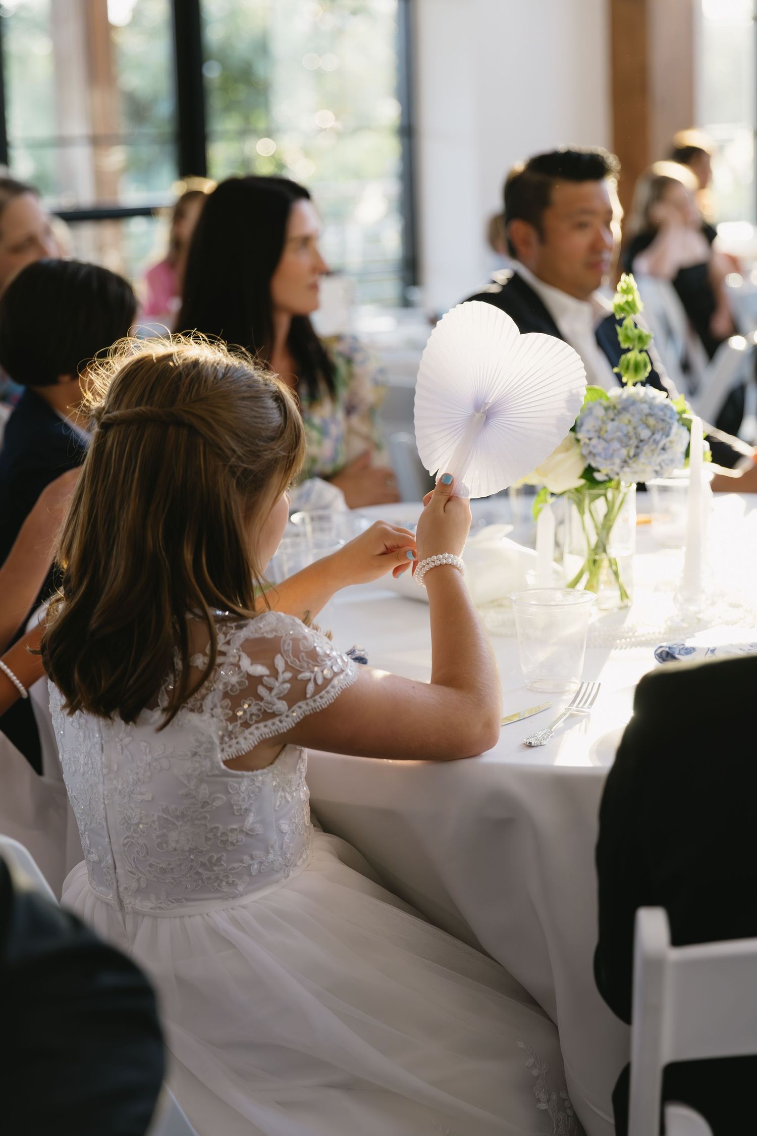 Wedding reception table setting with white flowers in vases and guests seated for dinner.
