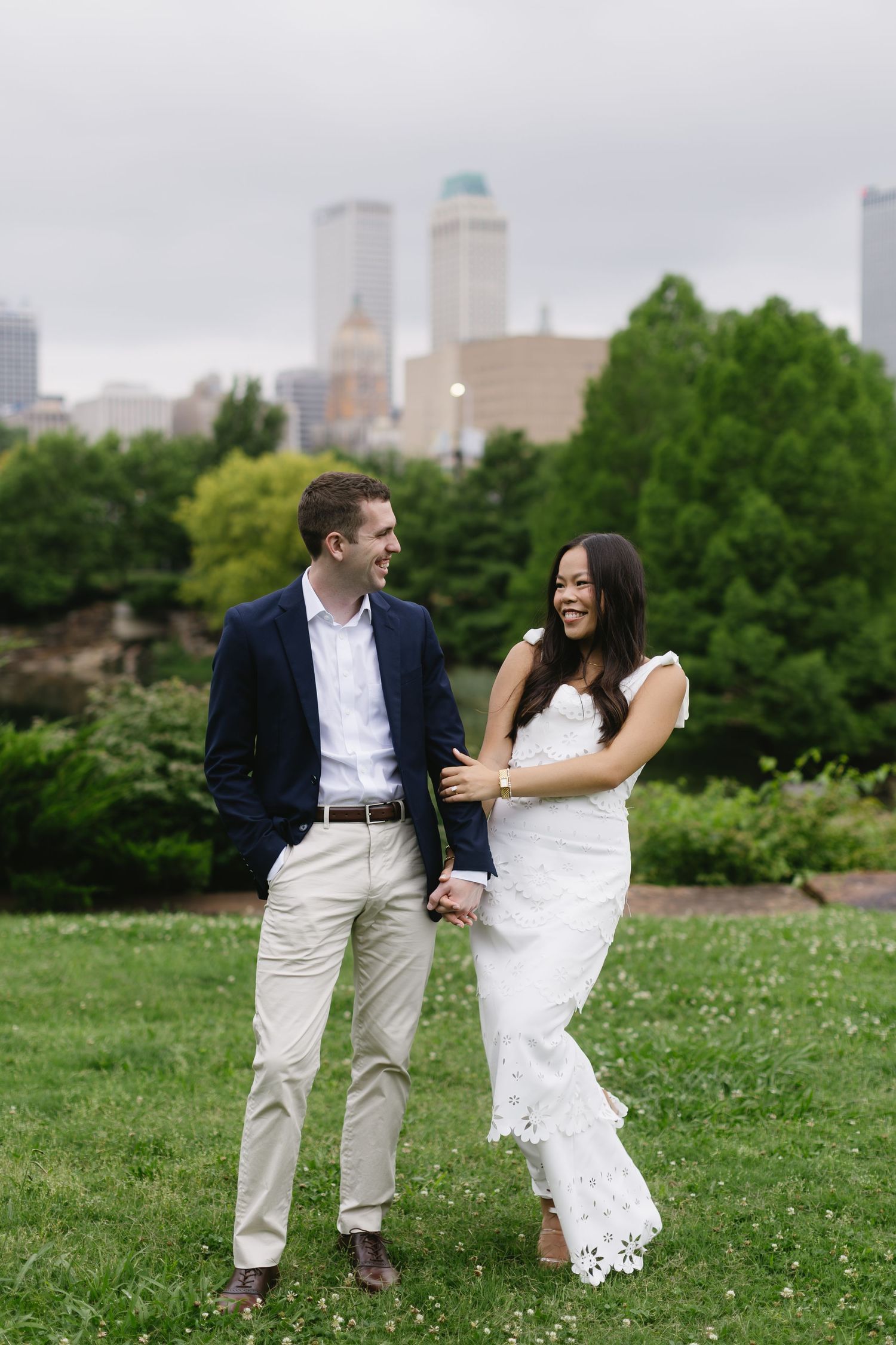 Couple shares playful moment in park with city skyline visible in background.