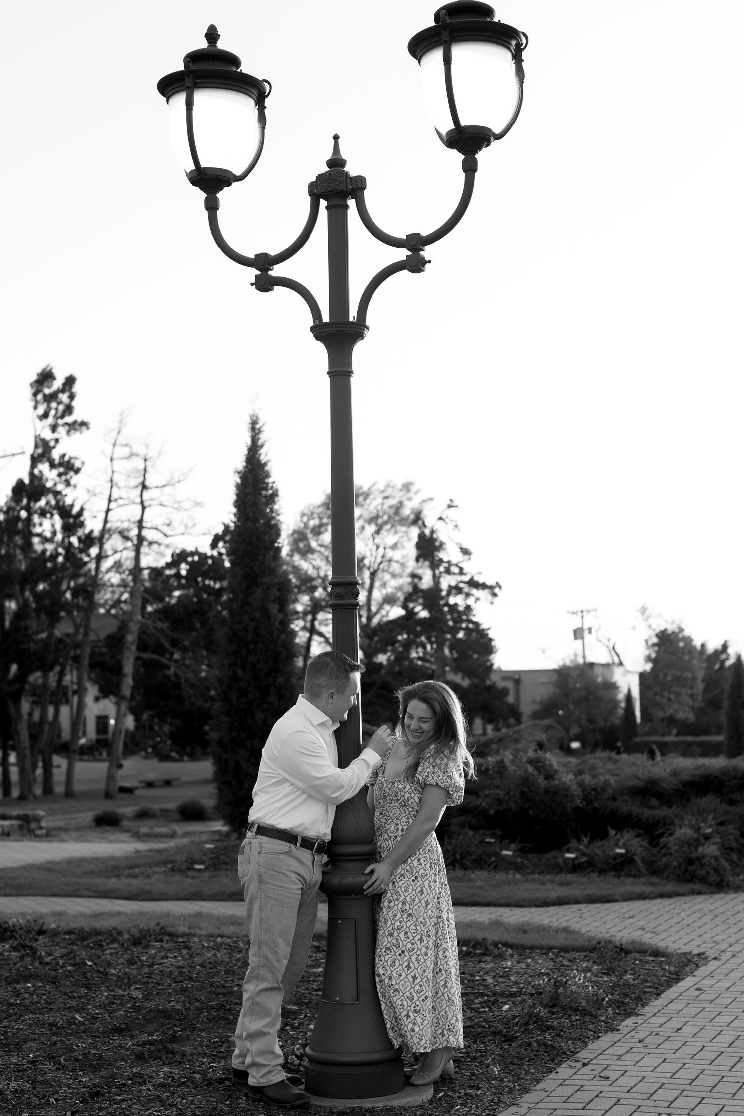 Black and white photo of couple posing playfully by vintage street lamps on brick path.