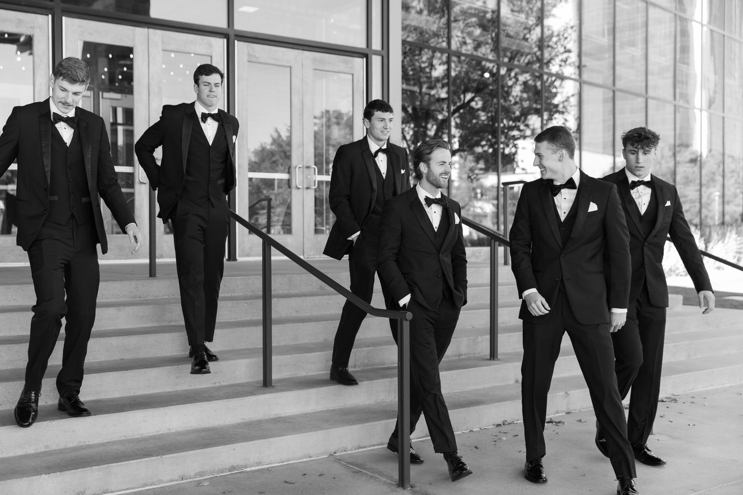 Groomsmen in black tuxedos pose on modern architectural steps in dramatic black and white.