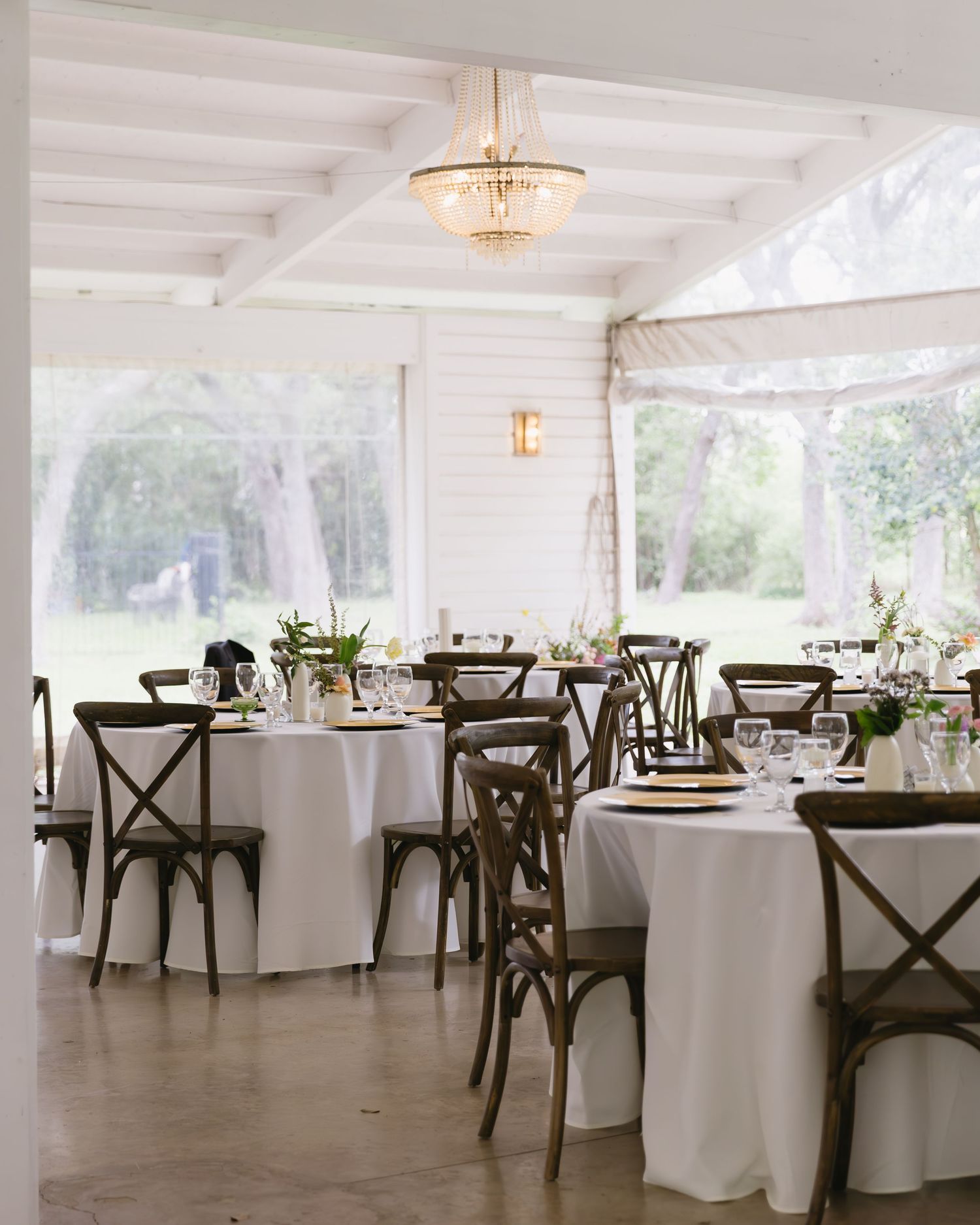 Elegant wedding reception setup with round tables, white tablecloths, and cross-back chairs in a bright white tent venue.
