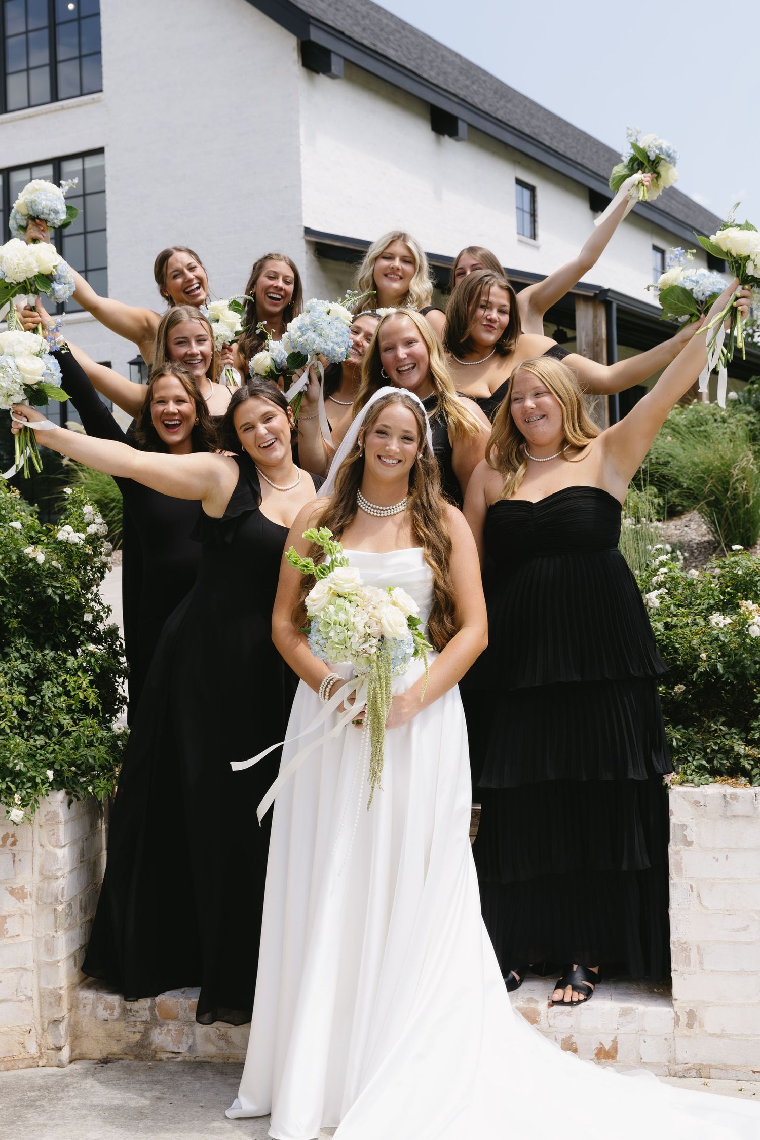 Wedding party poses joyfully outside with bridesmaids in black dresses and bride in white.