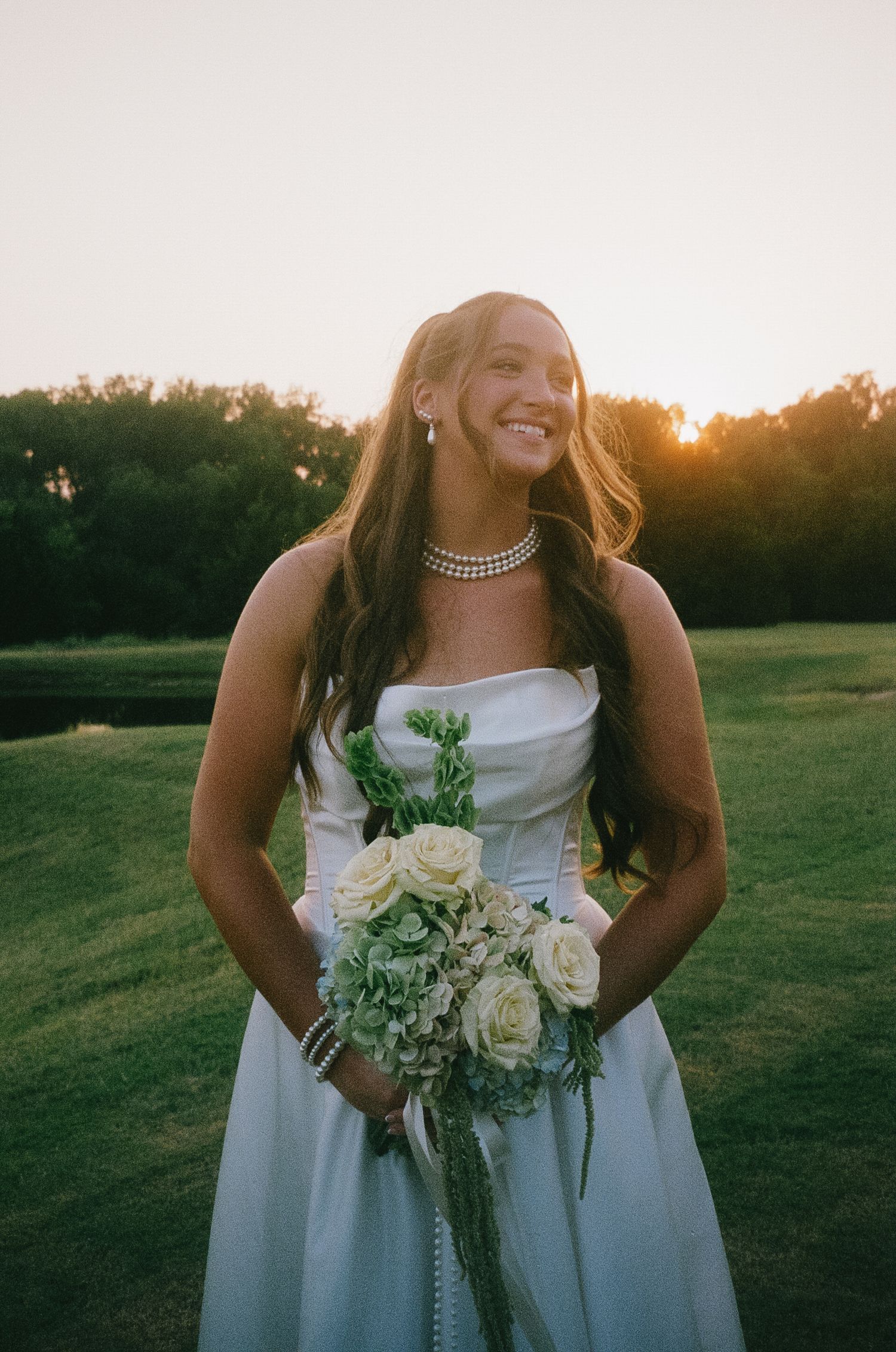 A bride in a white dress holds a bouquet of white roses at sunset in a grassy field.