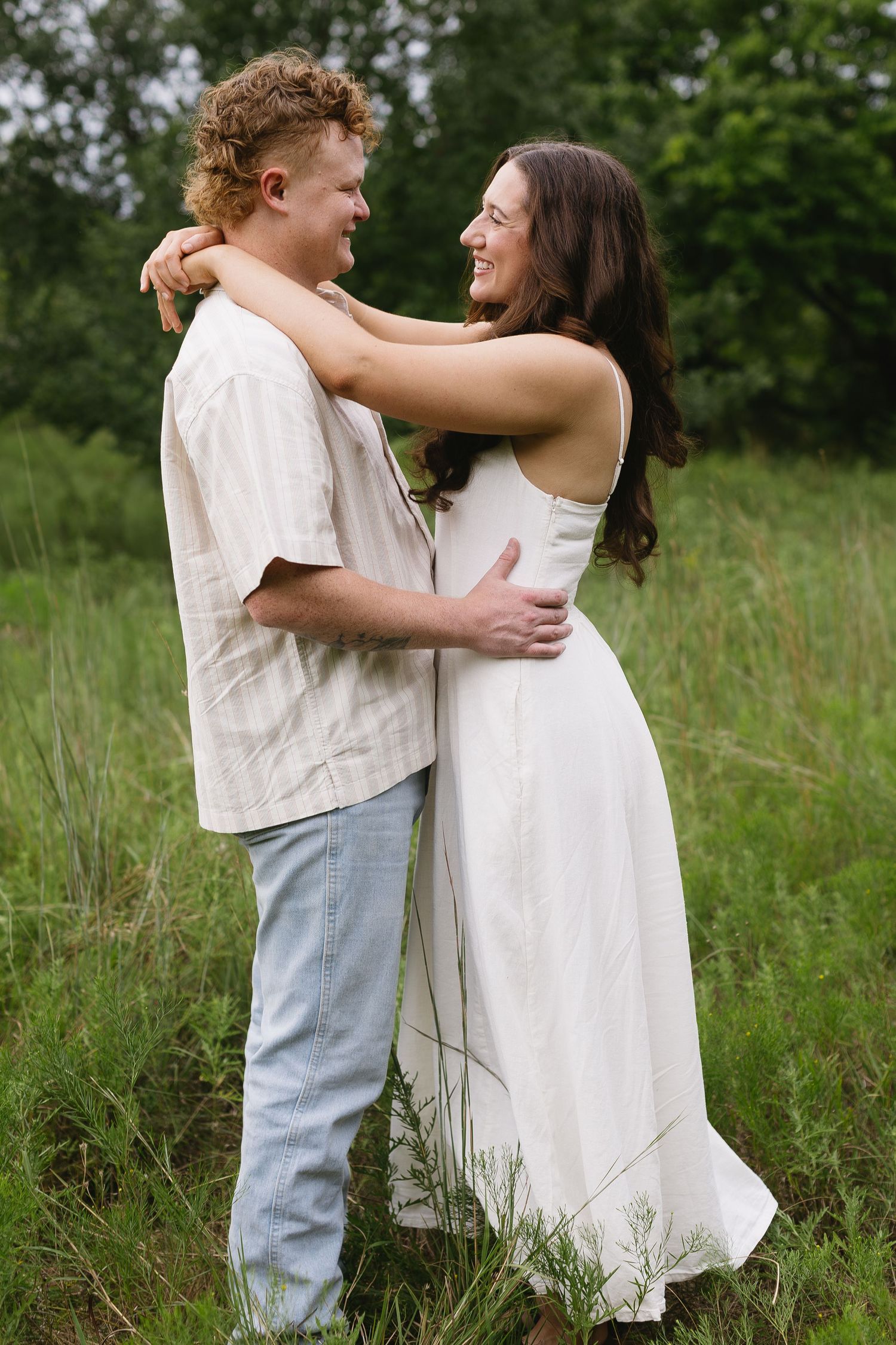 A couple in white clothing shares playful romantic moments while standing in a grassy meadow.