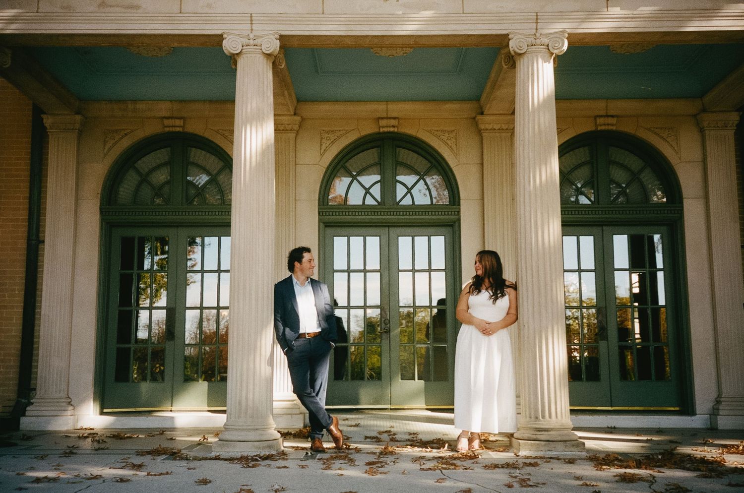Couple shares intimate moments in a grand colonnade hall with elegant white pillars and arched windows.