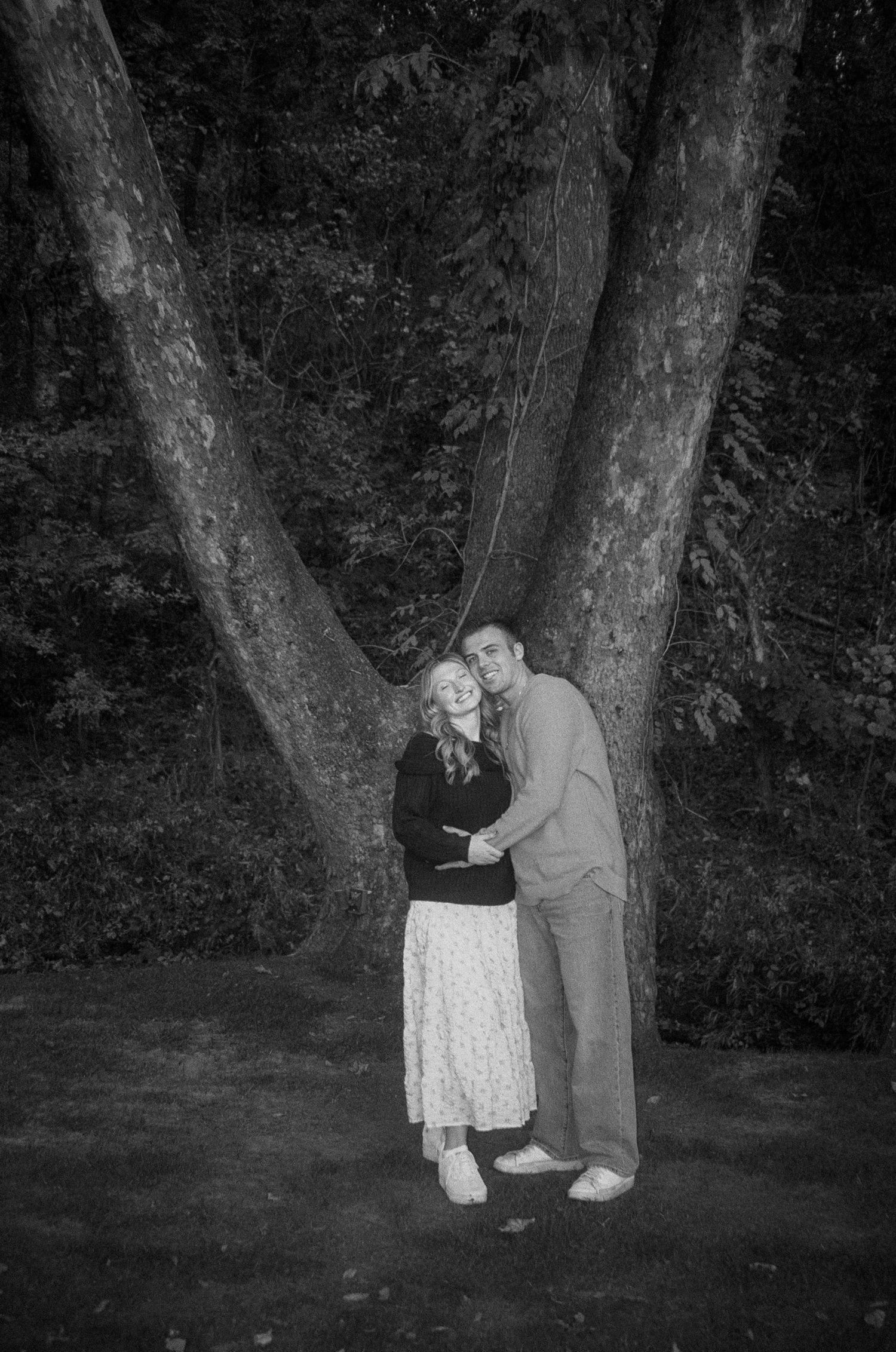 A black and white photo of a couple embracing by a distinctive V-shaped tree in a garden setting.
