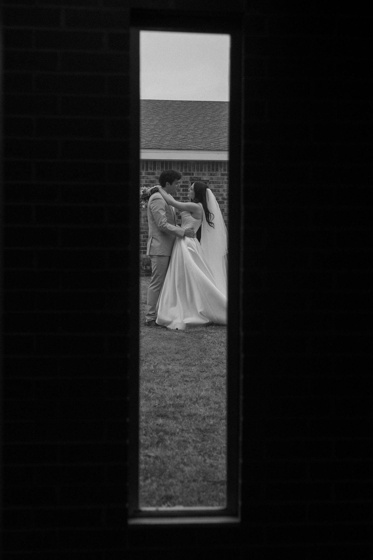 A black and white wedding photo framed through a narrow vertical window showing a couple dancing on grass.