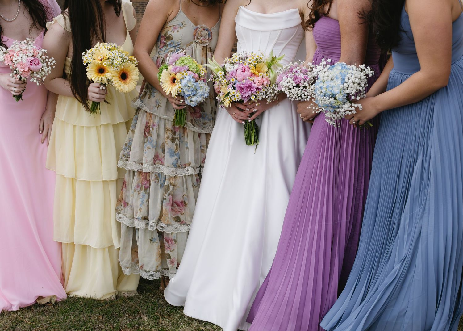 A row of bridesmaids in pastel colored dresses holding floral bouquets stand together at an outdoor wedding.