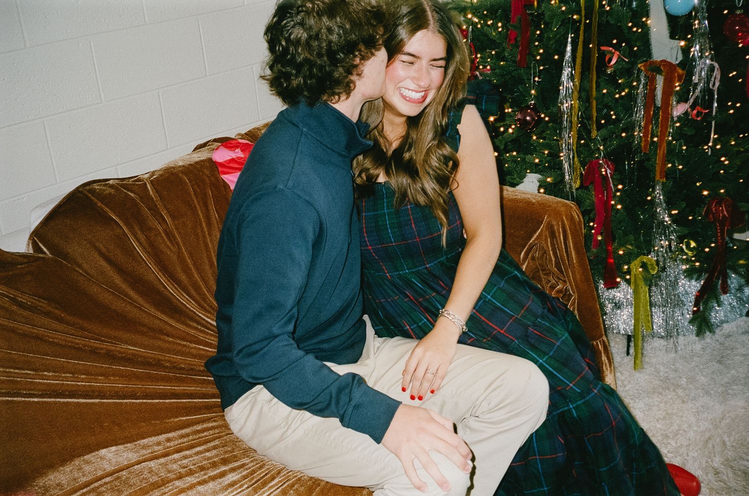 A couple in festive winter attire sits close together on a brown leather couch with a decorated Christmas tree in background.
