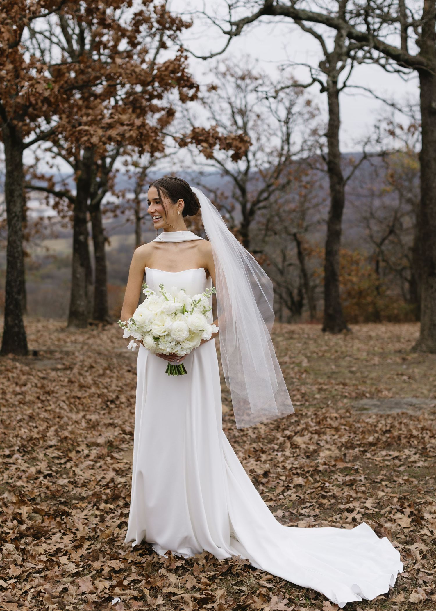 Bride in flowing white dress and veil stands among autumn trees holding a white bouquet.