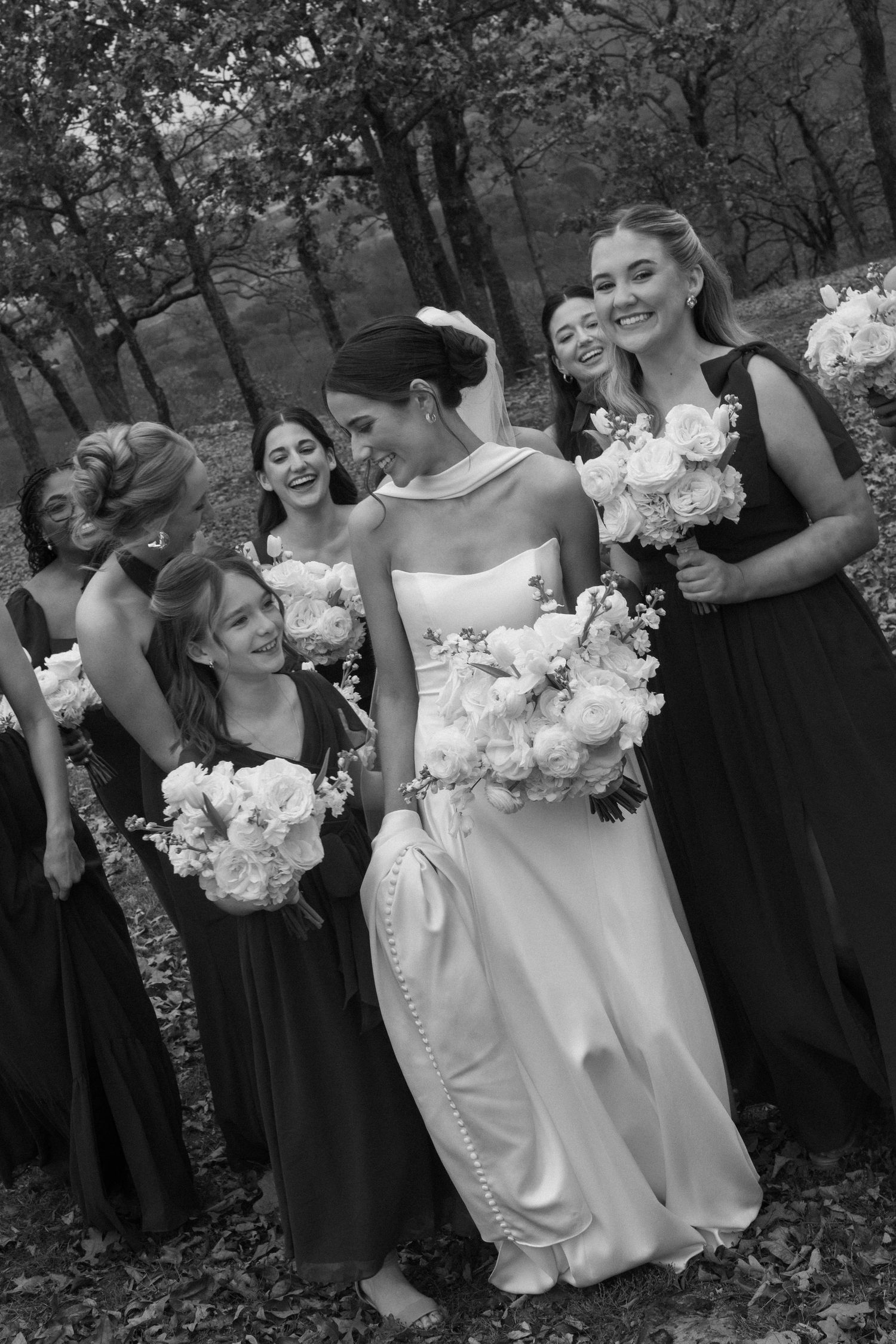 Bridesmaids in dark dresses pose with white floral bouquets in an outdoor garden setting.