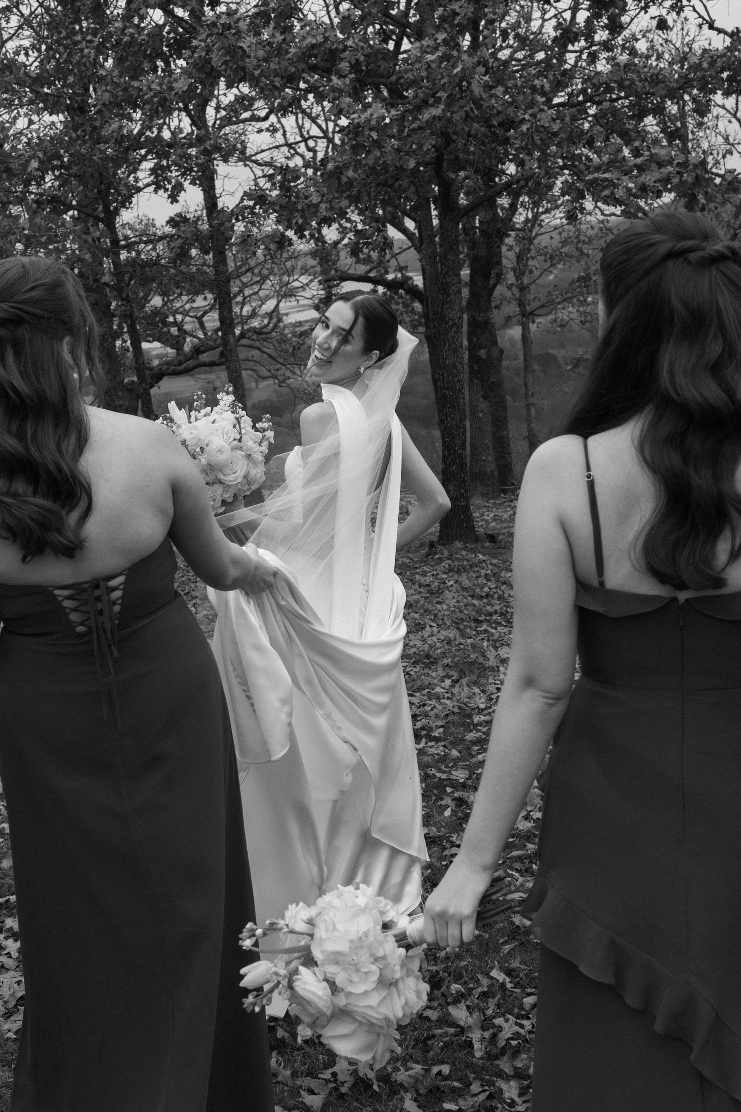Wedding party in black dresses and white gown gathered outdoors among autumn trees.