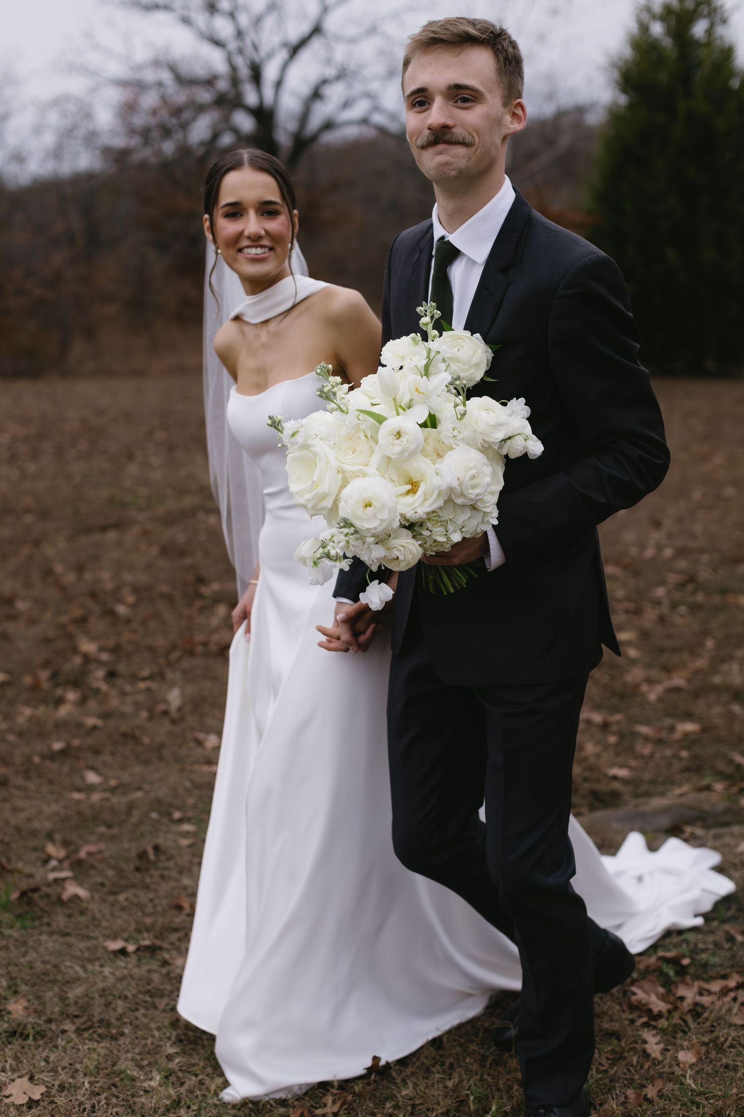Wedding couple posing in outdoor fall setting with white bouquet and elegant attire.