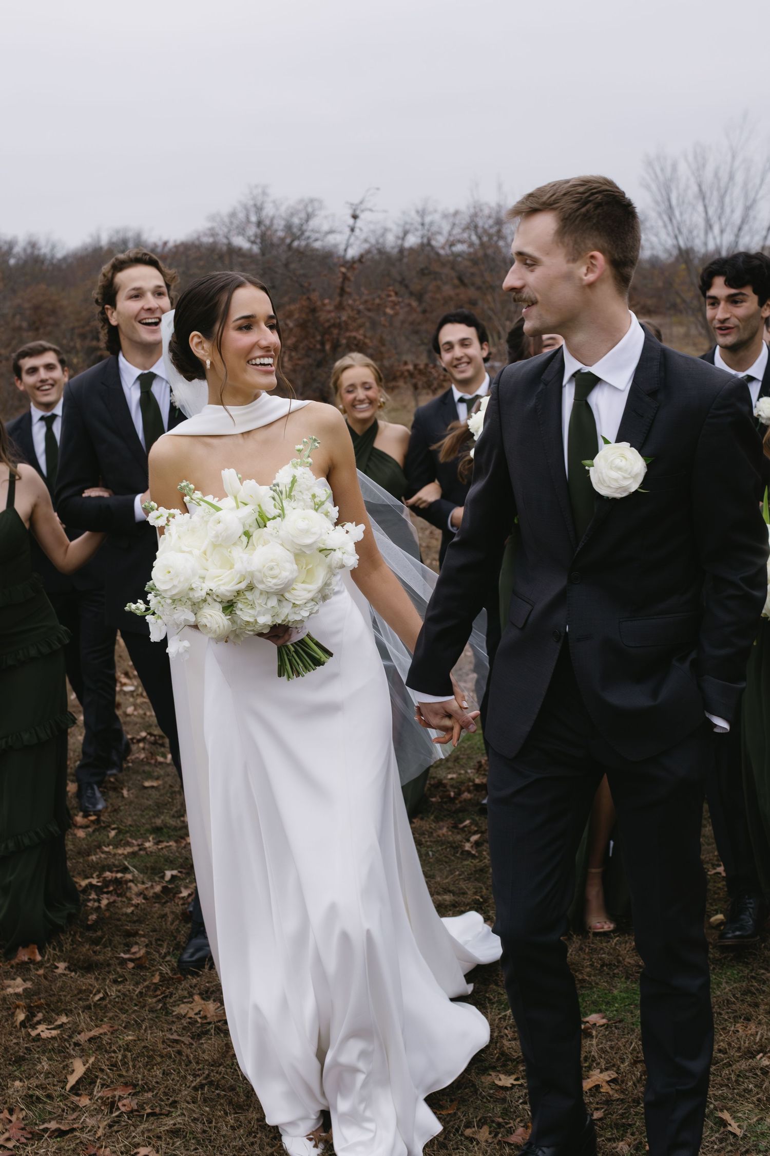 Wedding party in formal attire gathers outdoors for autumn ceremony photos.