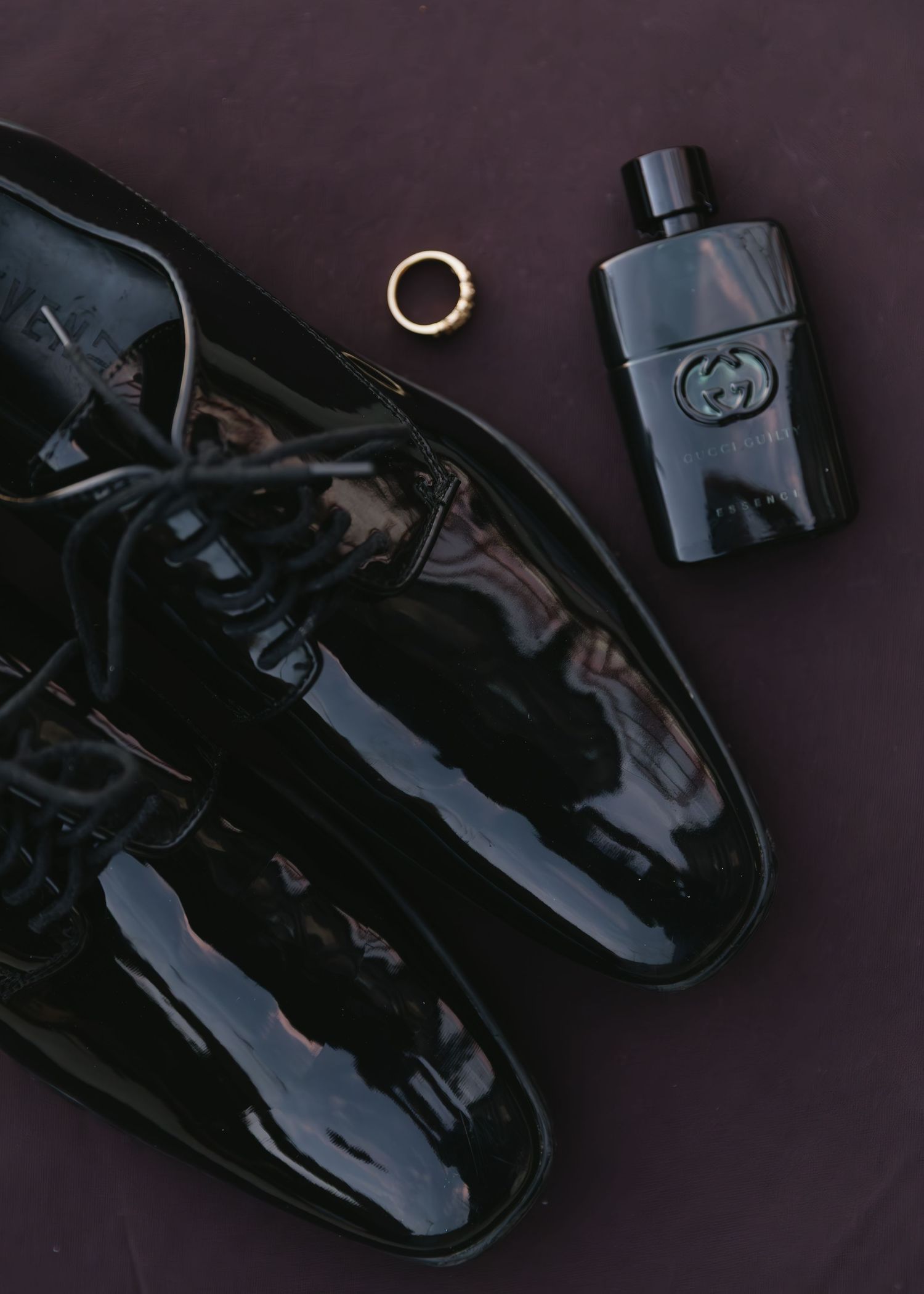 Wedding accessories including patent leather shoes, gold ring and Gucci cologne on dark surface.