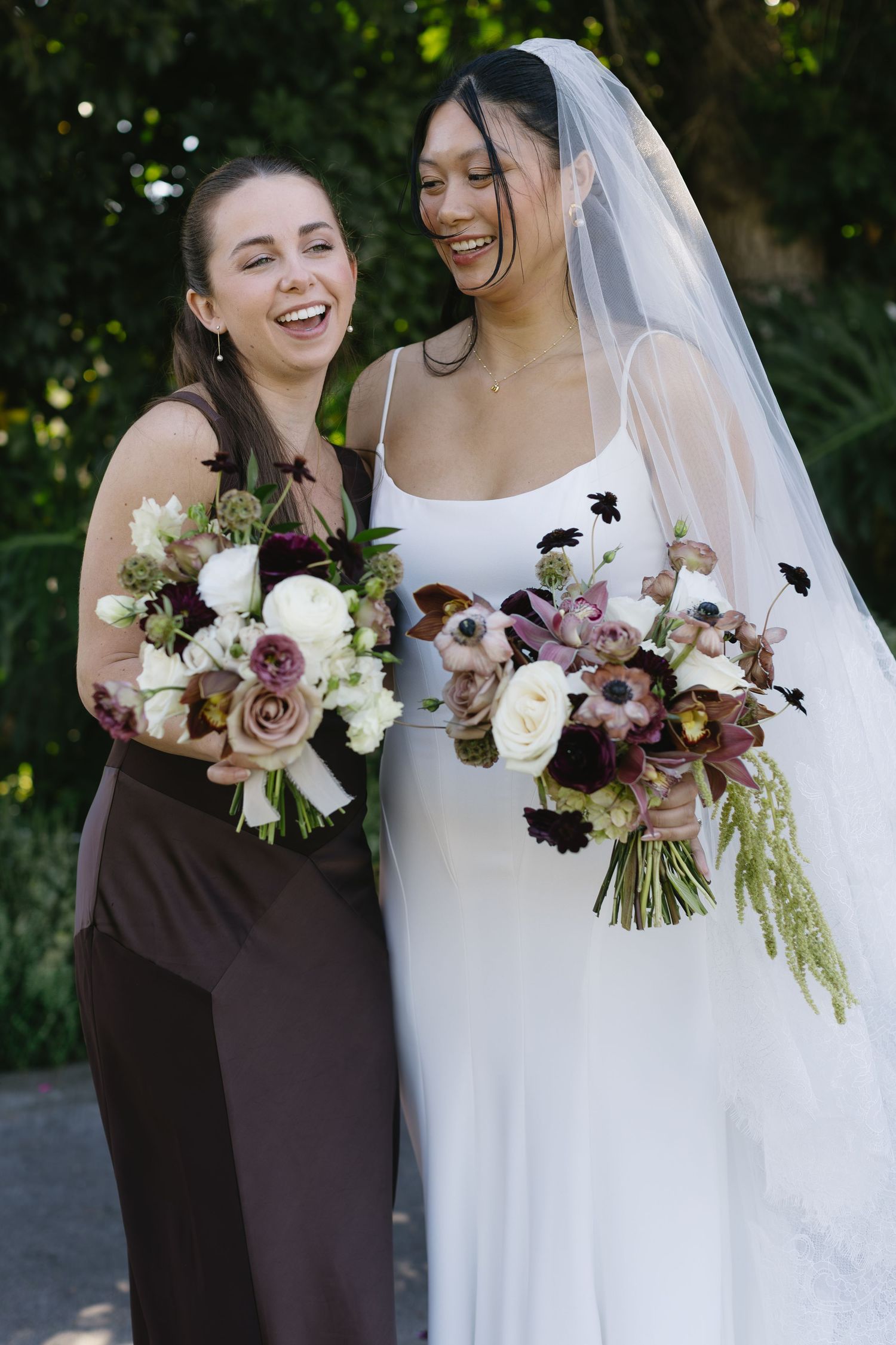 Bride and bridesmaid share genuine laughter while holding matching floral bouquets.