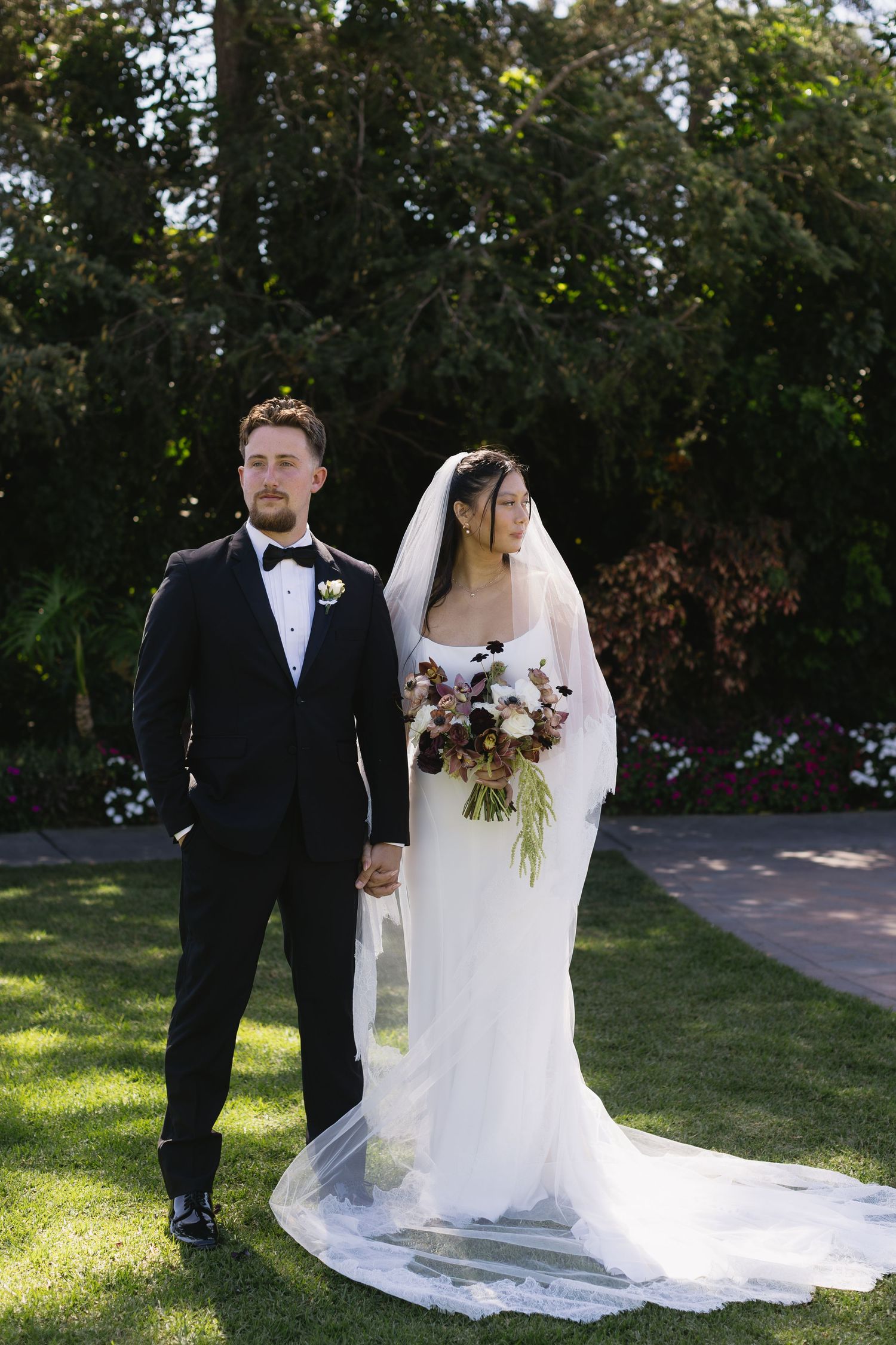 Newlyweds pose in garden setting with bride's long train flowing behind.
