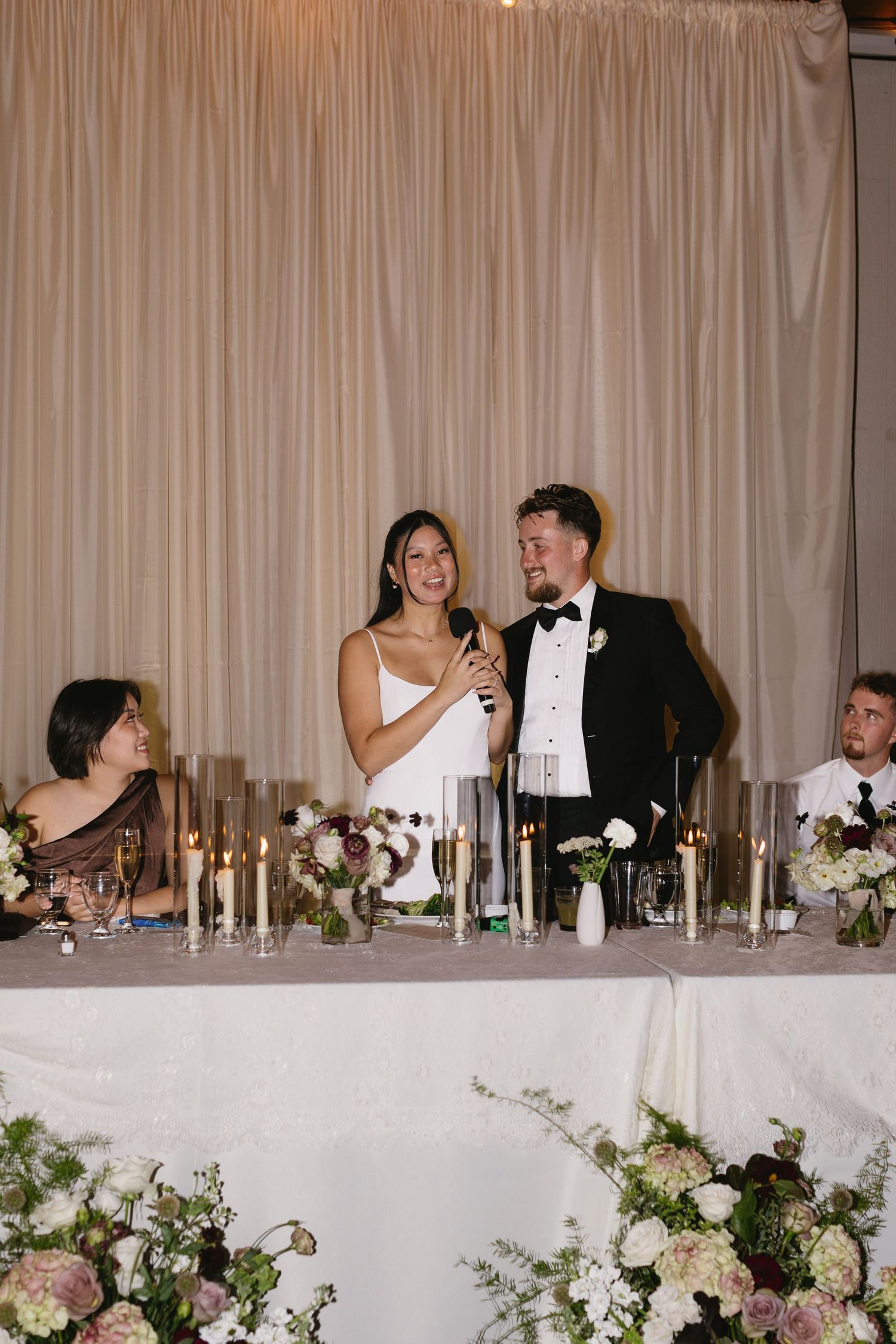 Wedding party celebrates at elegant head table with white floral decorations and draped backdrop.