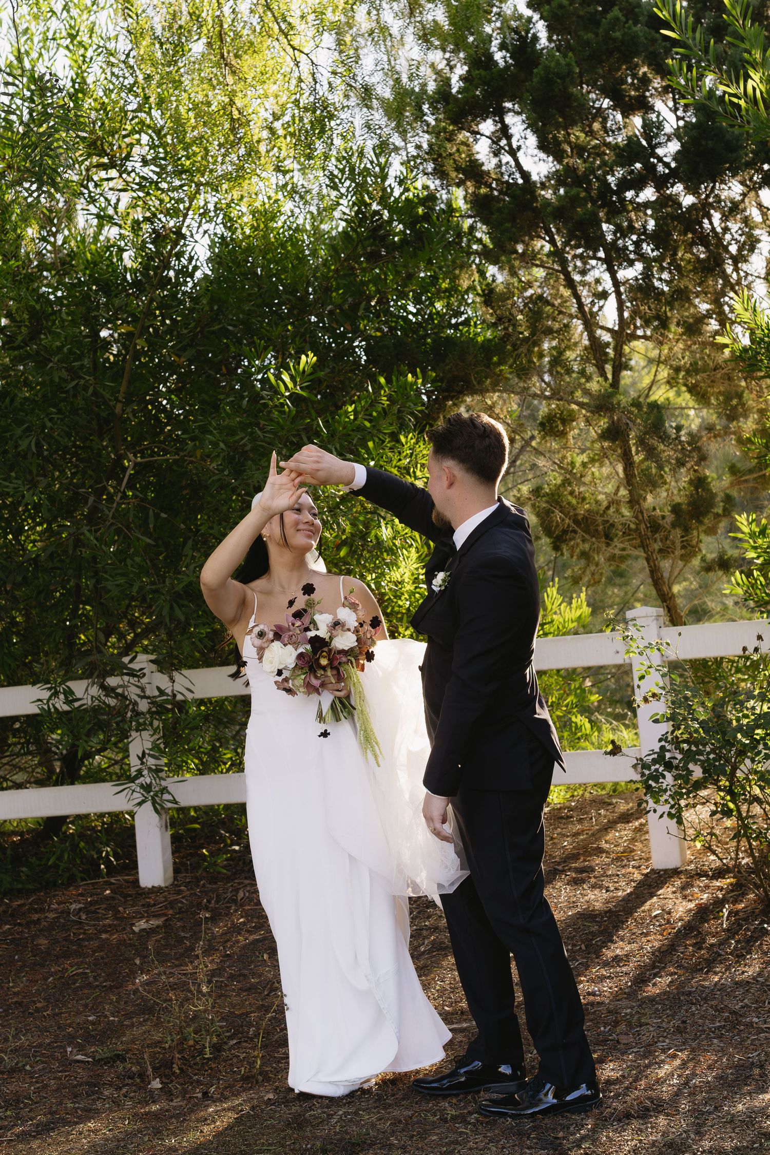 Bride and groom share playful moments along a white fence in an outdoor garden setting.