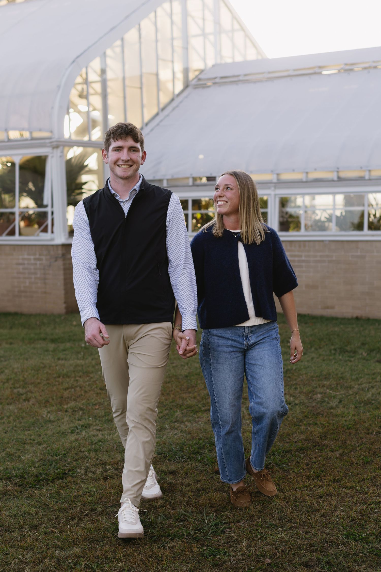 A couple walks together on grass in front of a white building during golden hour.