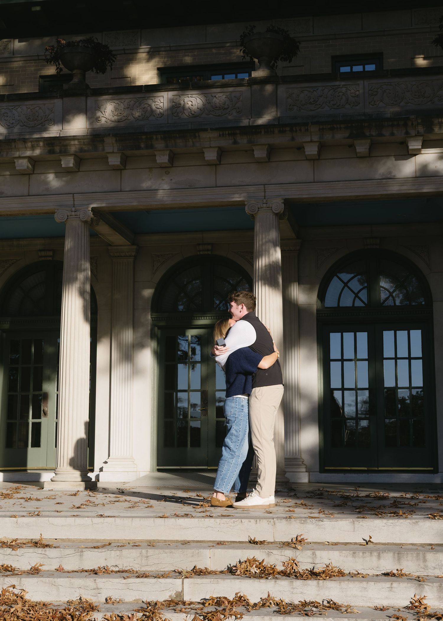 A couple embracing near classical columns with fallen leaves.