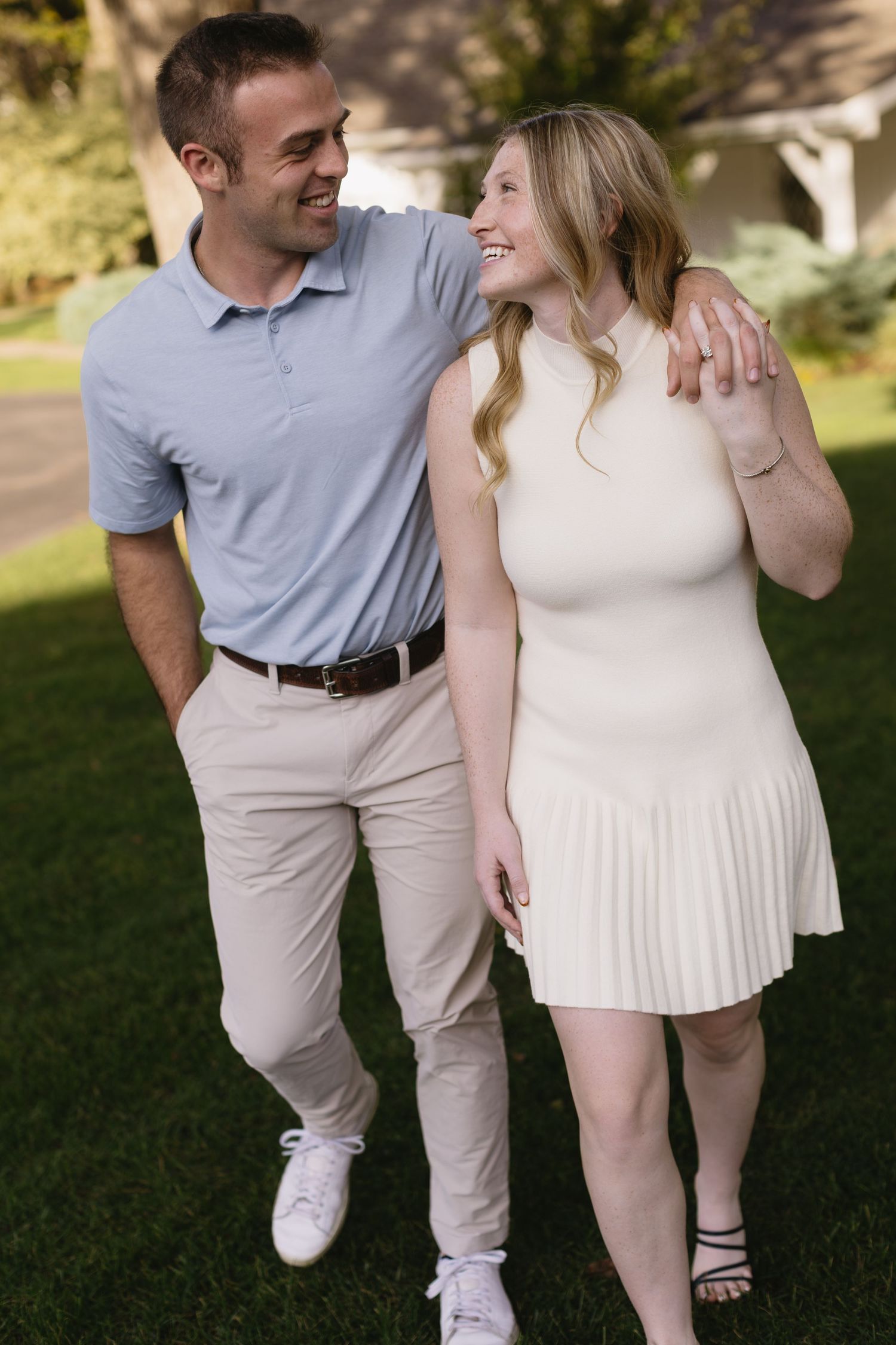 A couple in light summer attire walks together on lush green grass during an outdoor photoshoot.