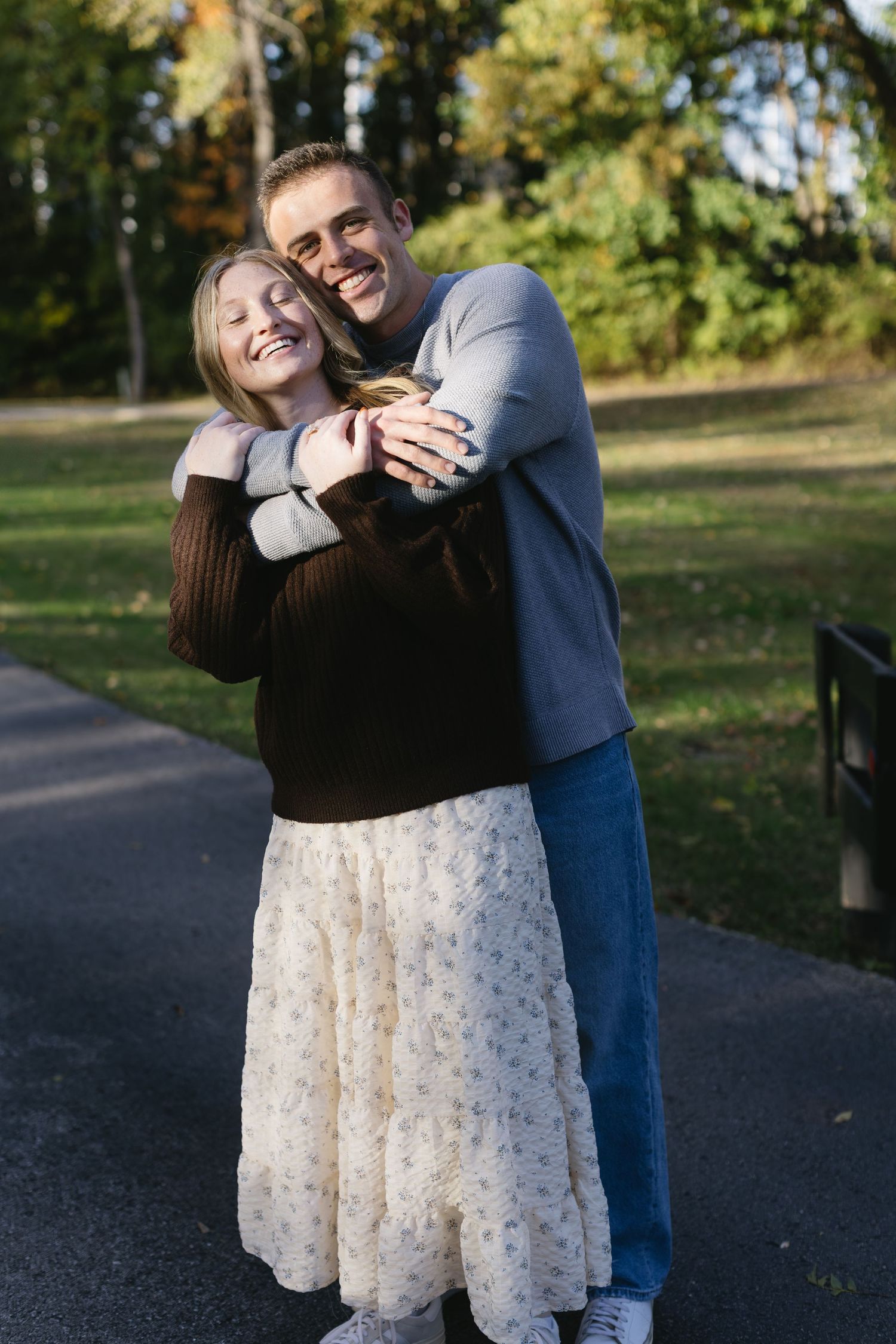 A couple embraces on a park path wearing casual fall attire with trees in the background.