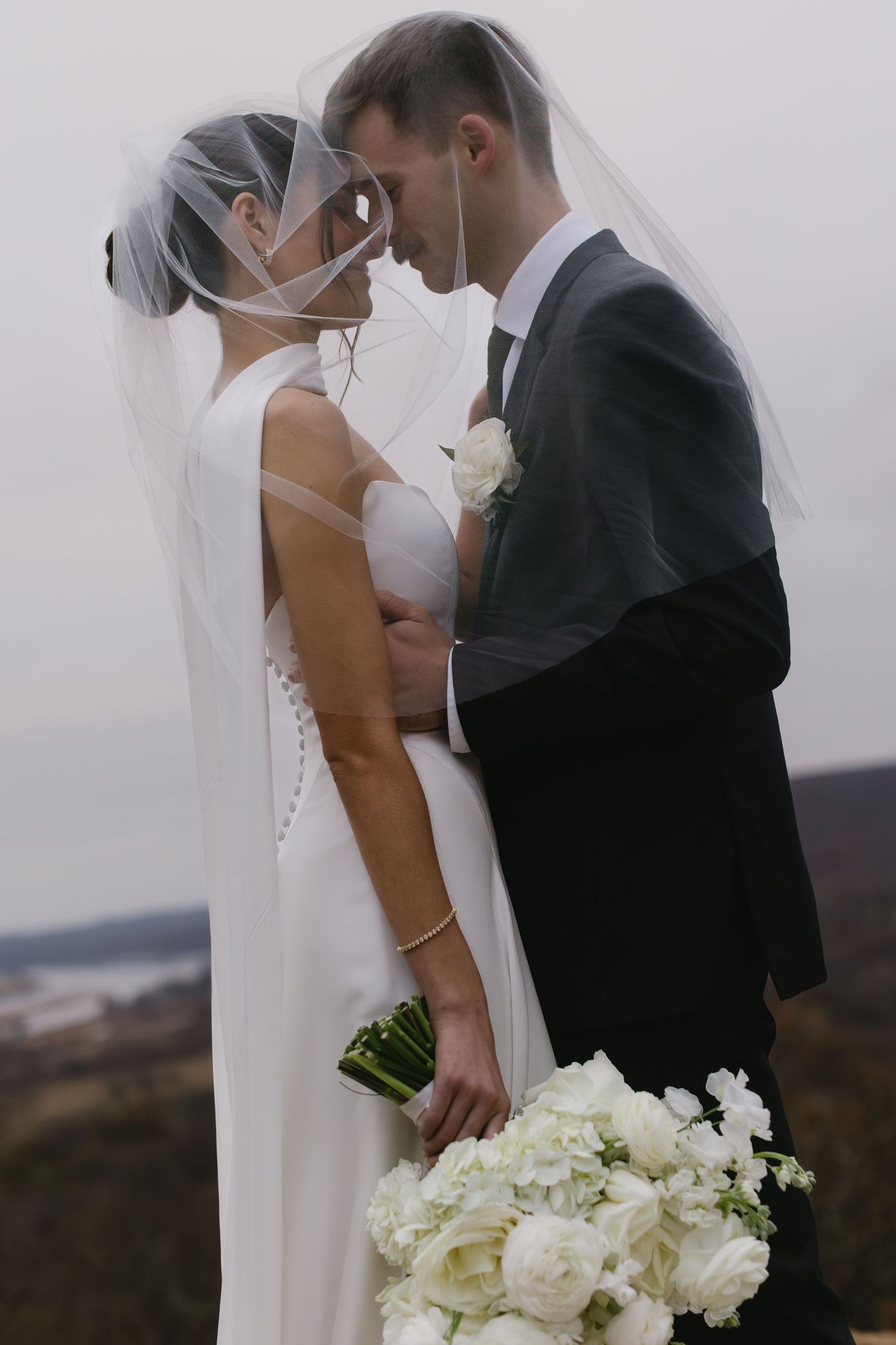 A romantic wedding couple shares an intimate kiss while holding a white floral bouquet against a misty outdoor backdrop.