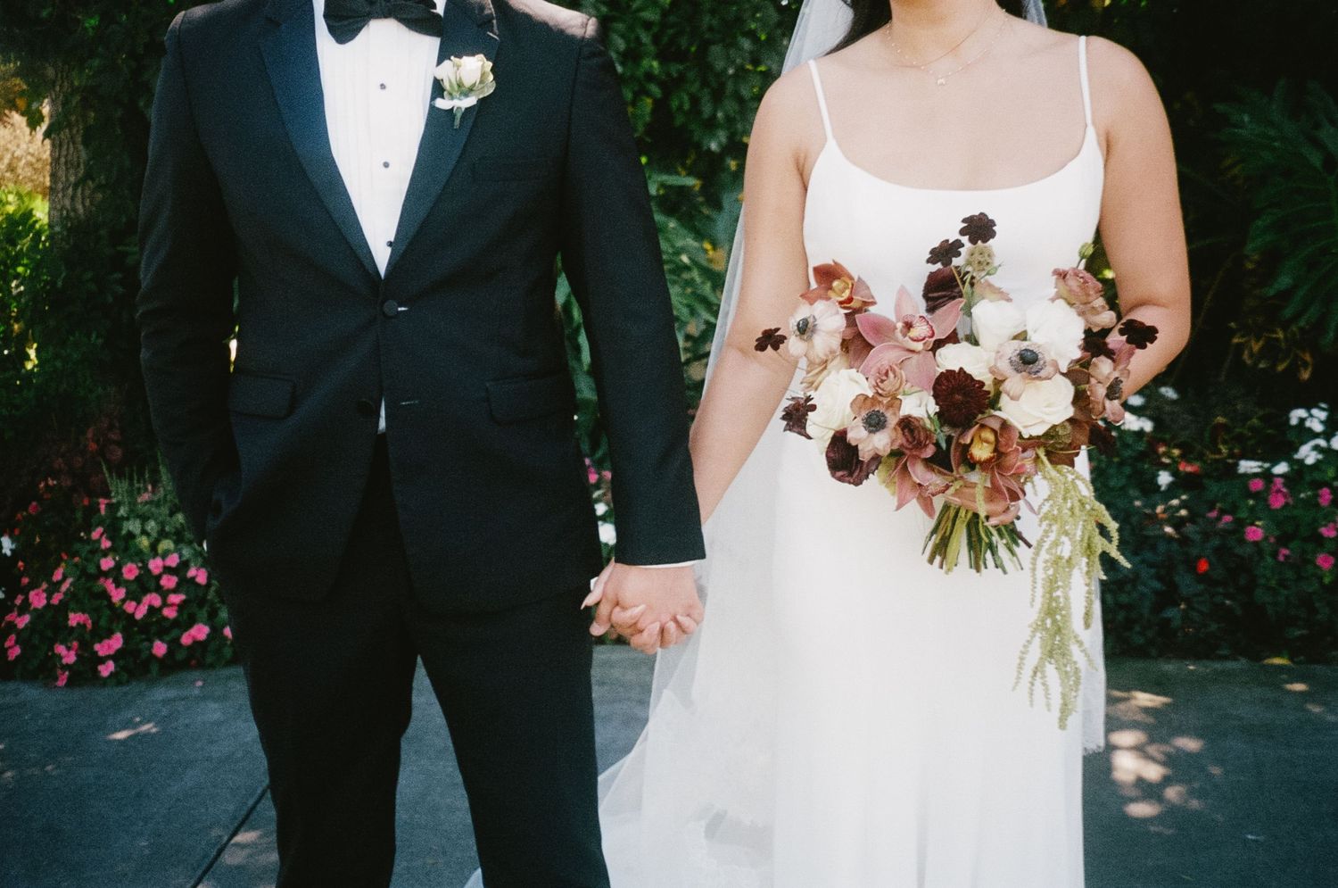 Wedding couple in formal attire standing together with floral bouquet in a garden setting.